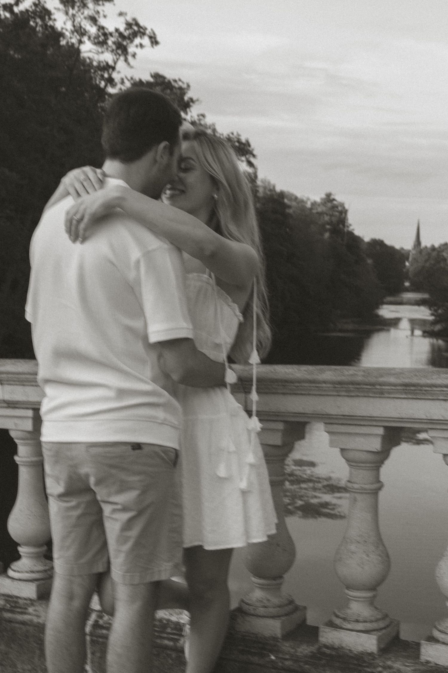 A couple in white outfits shares an intimate moment by a stone balustrade overlooking water.