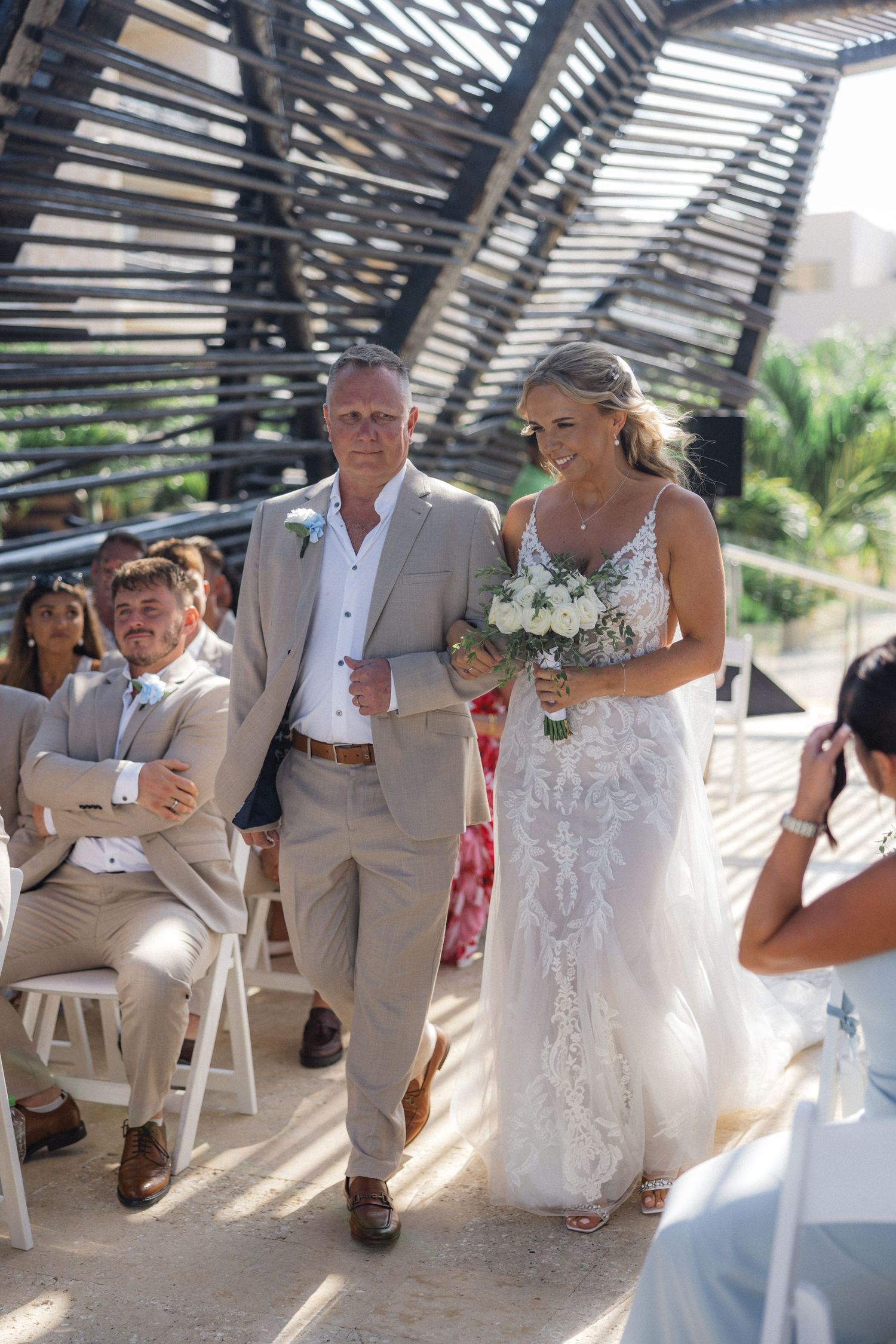 Wedding ceremony under pergola with bride in white lace dress and guests in formal attire.