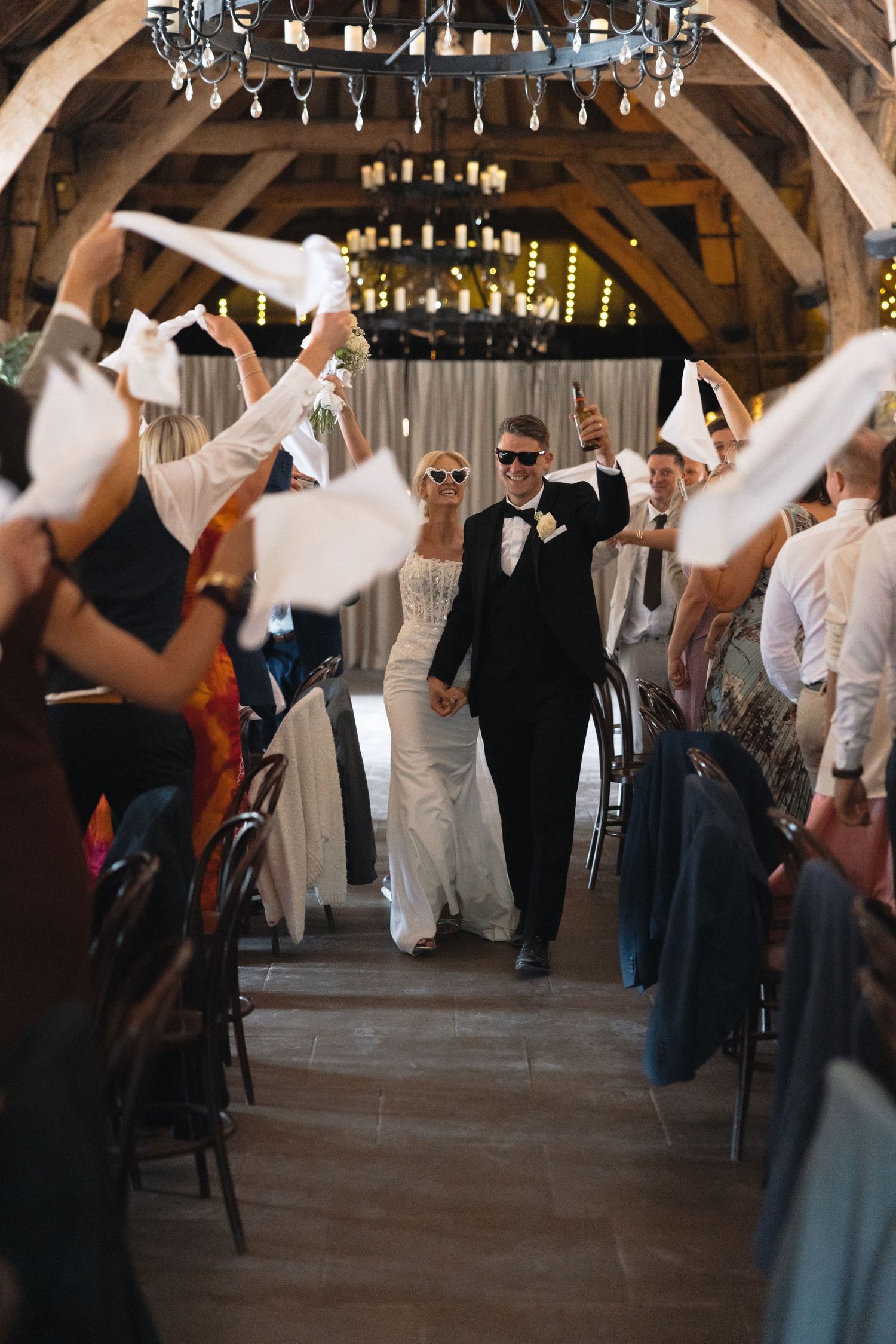 Wedding celebration with guests waving napkins as couple walks through rustic venue.