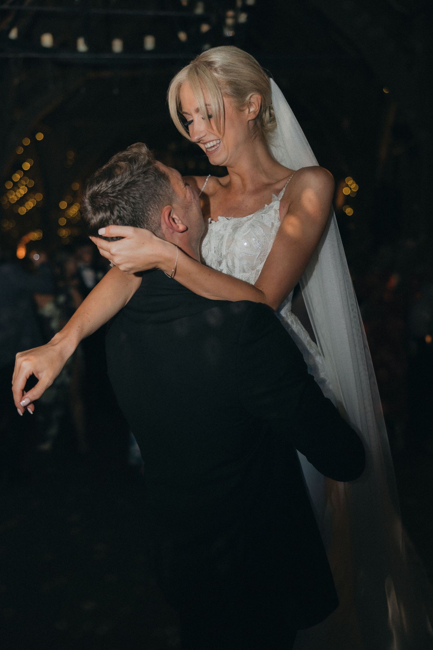 A bride in white dress and veil leans over her dancing partner during a wedding celebration.