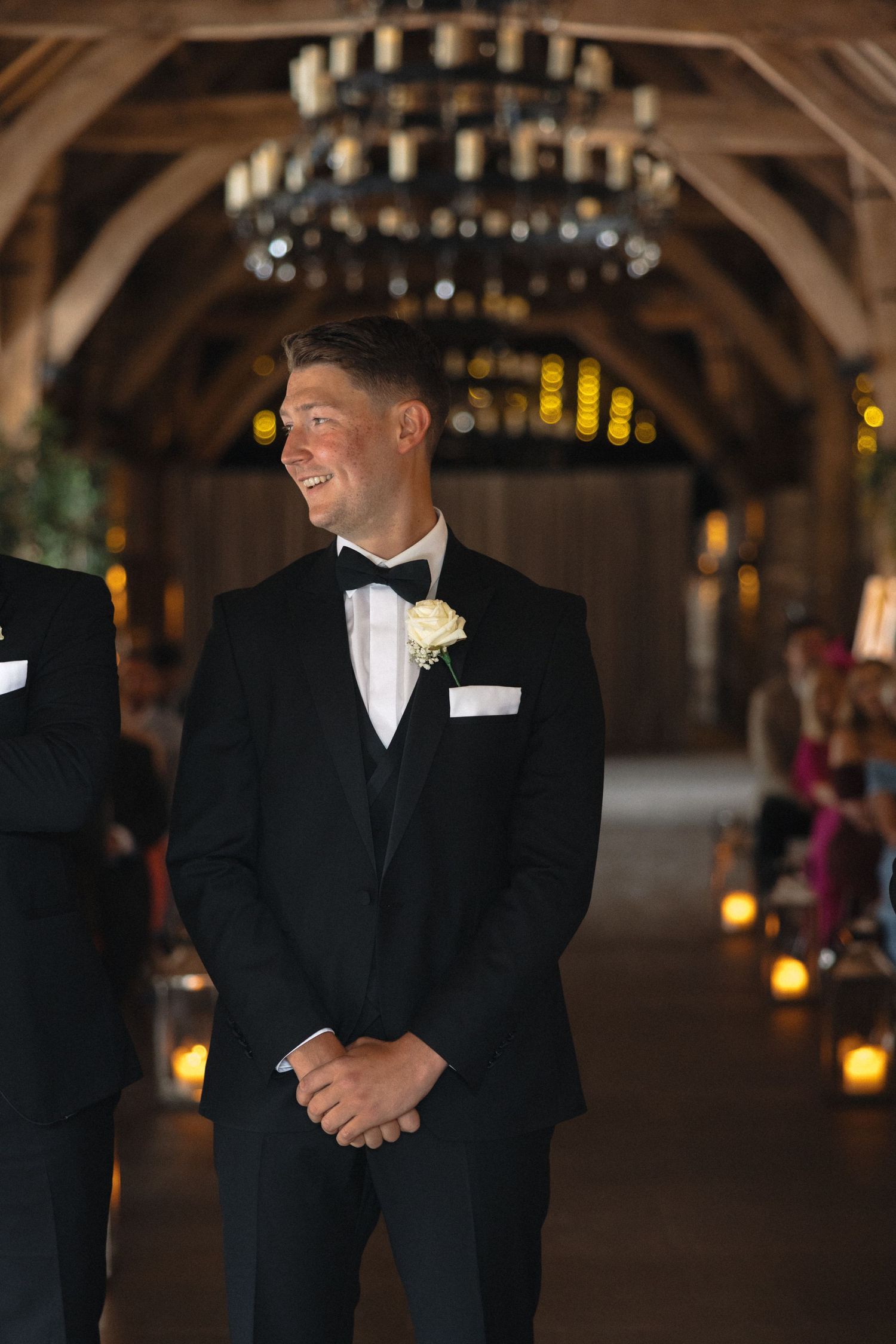 Groomsmen in black tuxedos stand in a rustic venue with wooden beams and chandeliers.