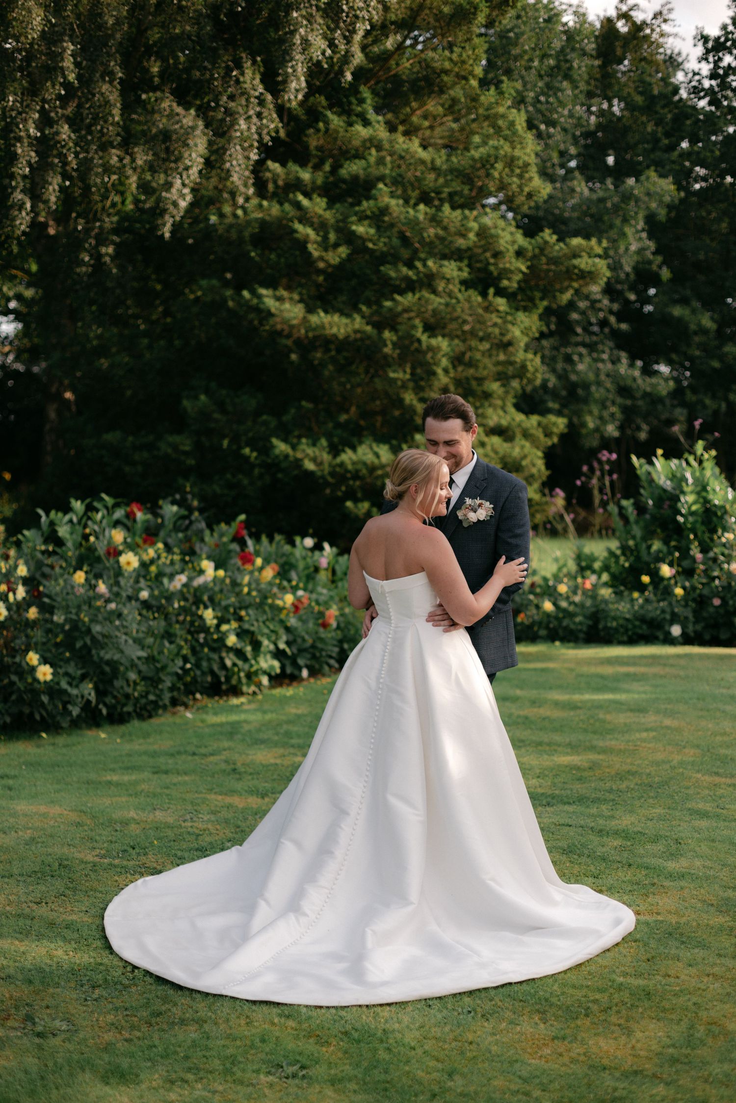 A wedding couple embraces on a lush green lawn with flowers in the background.
