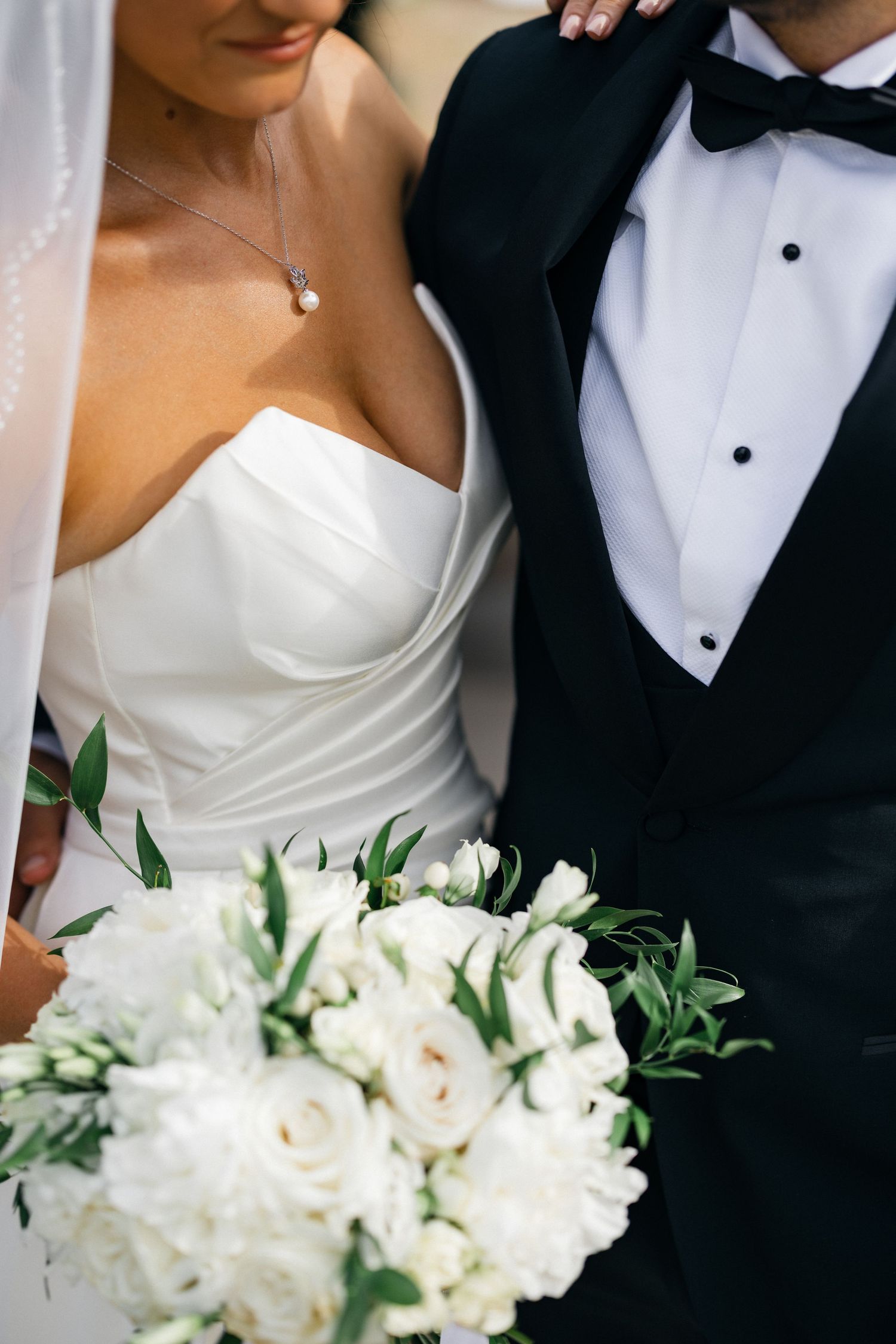 Close up details of a bride in white dress and groom in tuxedo with white floral bouquet.