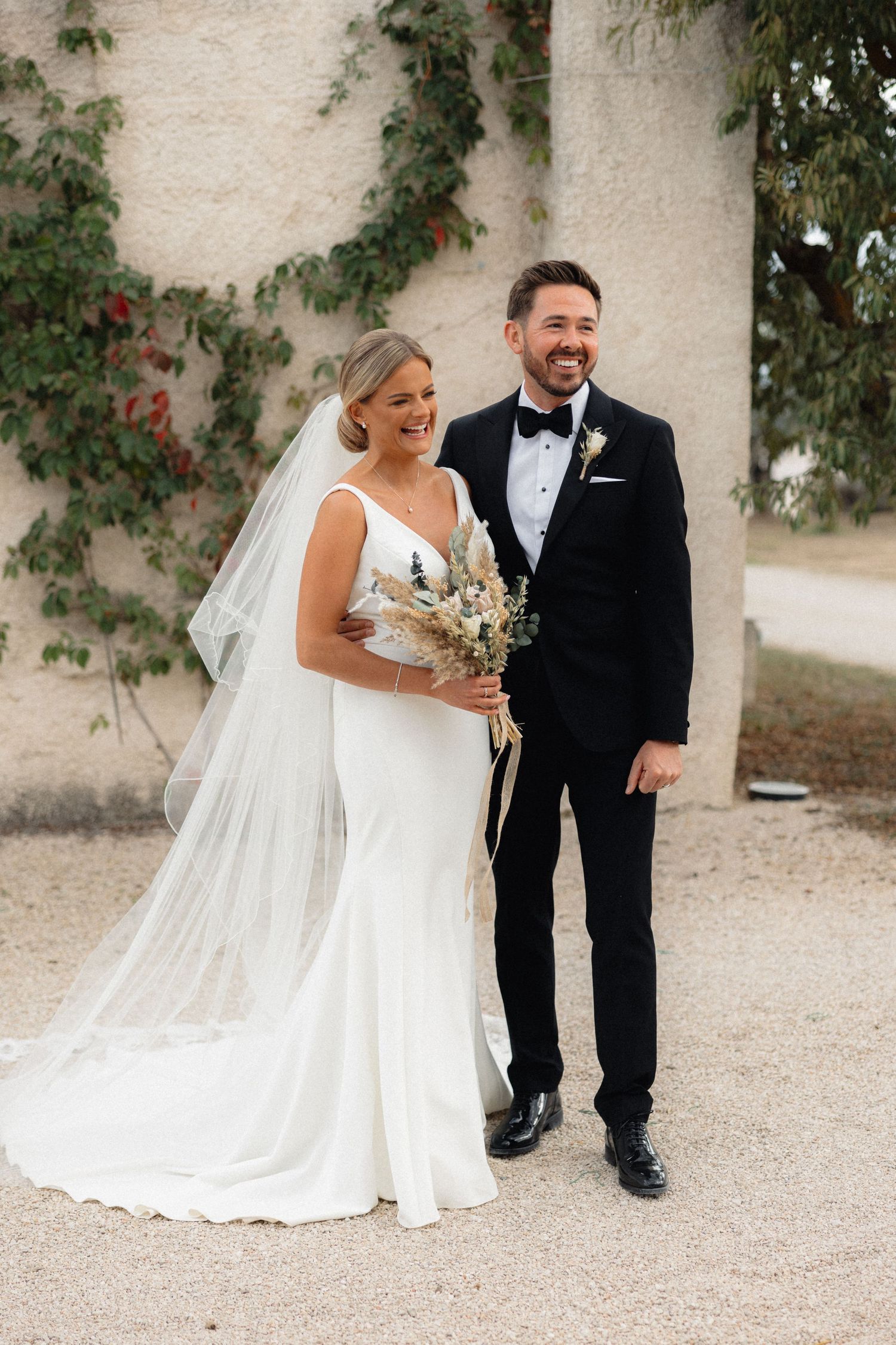 Candid wedding couple laughing together against white wall with climbing vines.