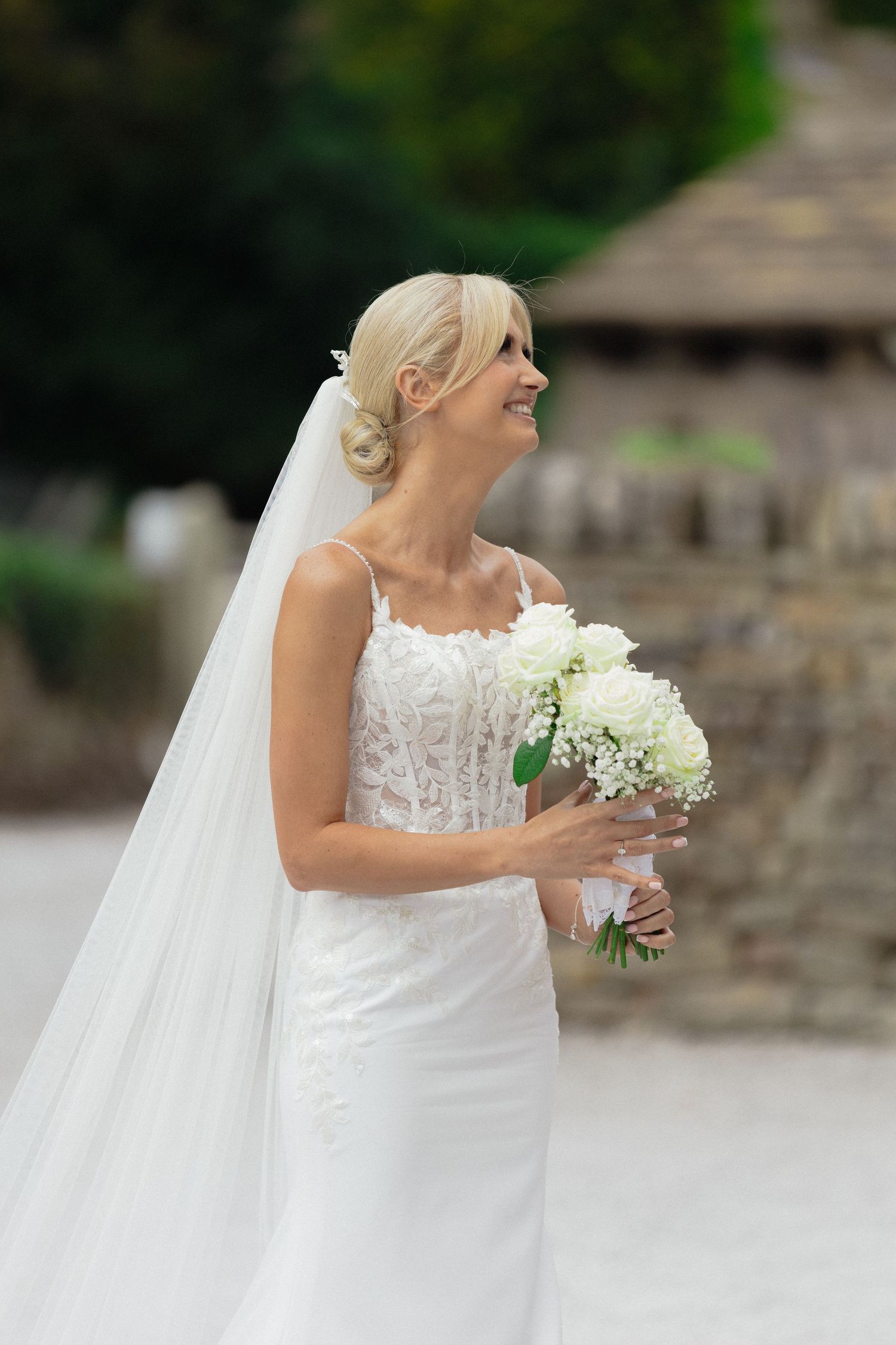 A bride in a lace wedding dress holds a white floral bouquet against a stone wall backdrop.