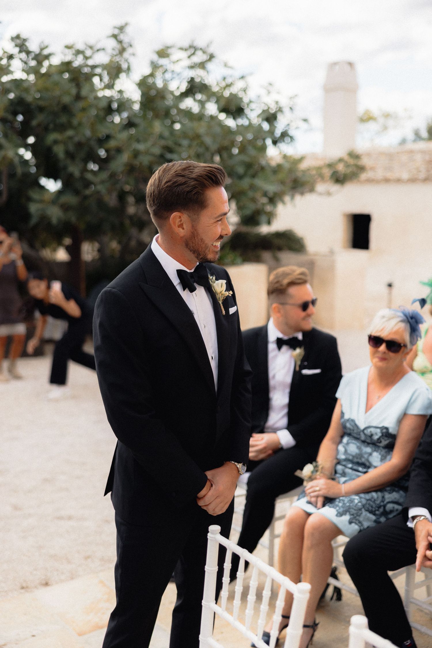 Wedding party members interact with guests seated in white chairs during an outdoor ceremony.