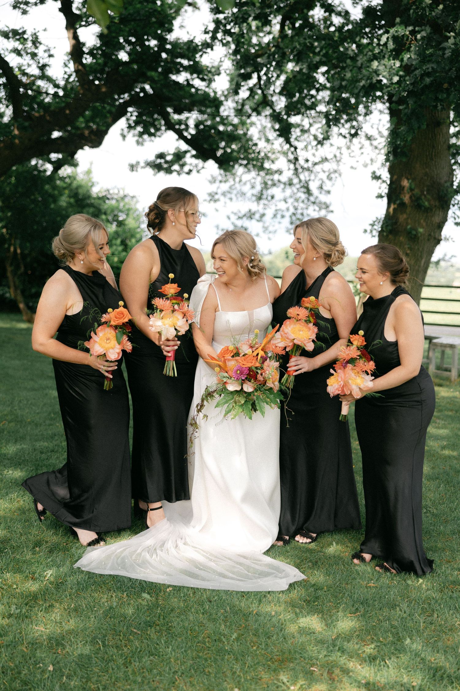 Bride in white dress poses with bridesmaids in black dresses holding orange bouquets.