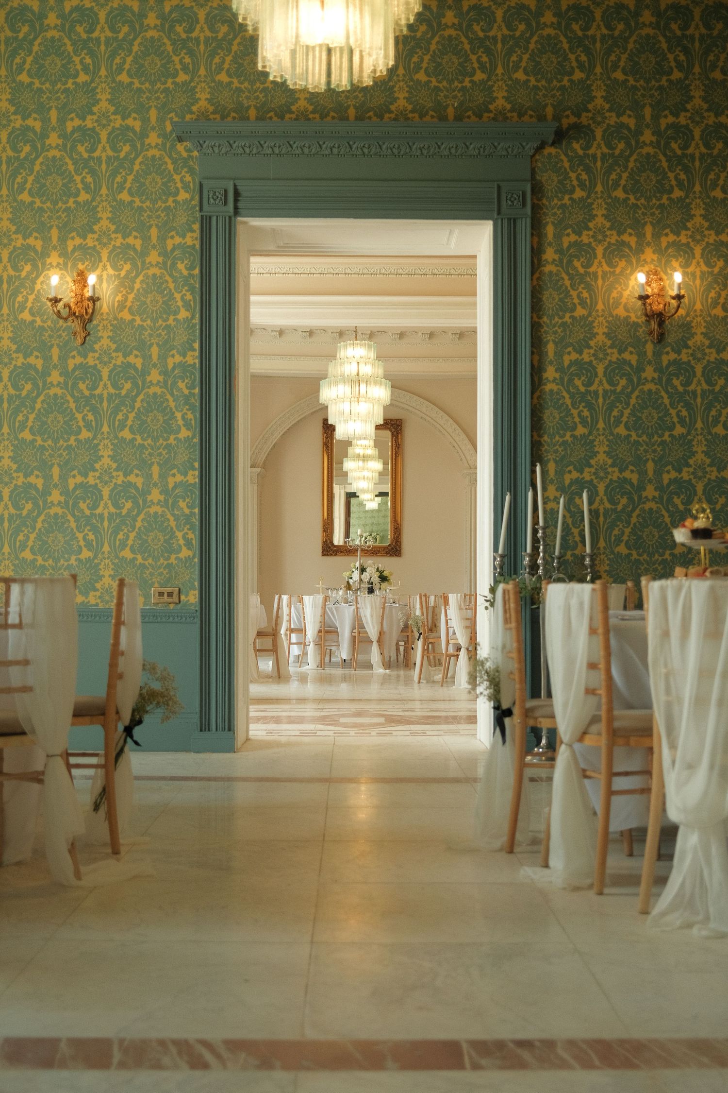 Elegant ballroom with teal damask wallpaper, crystal chandeliers, and white-draped chairs for a wedding reception.
