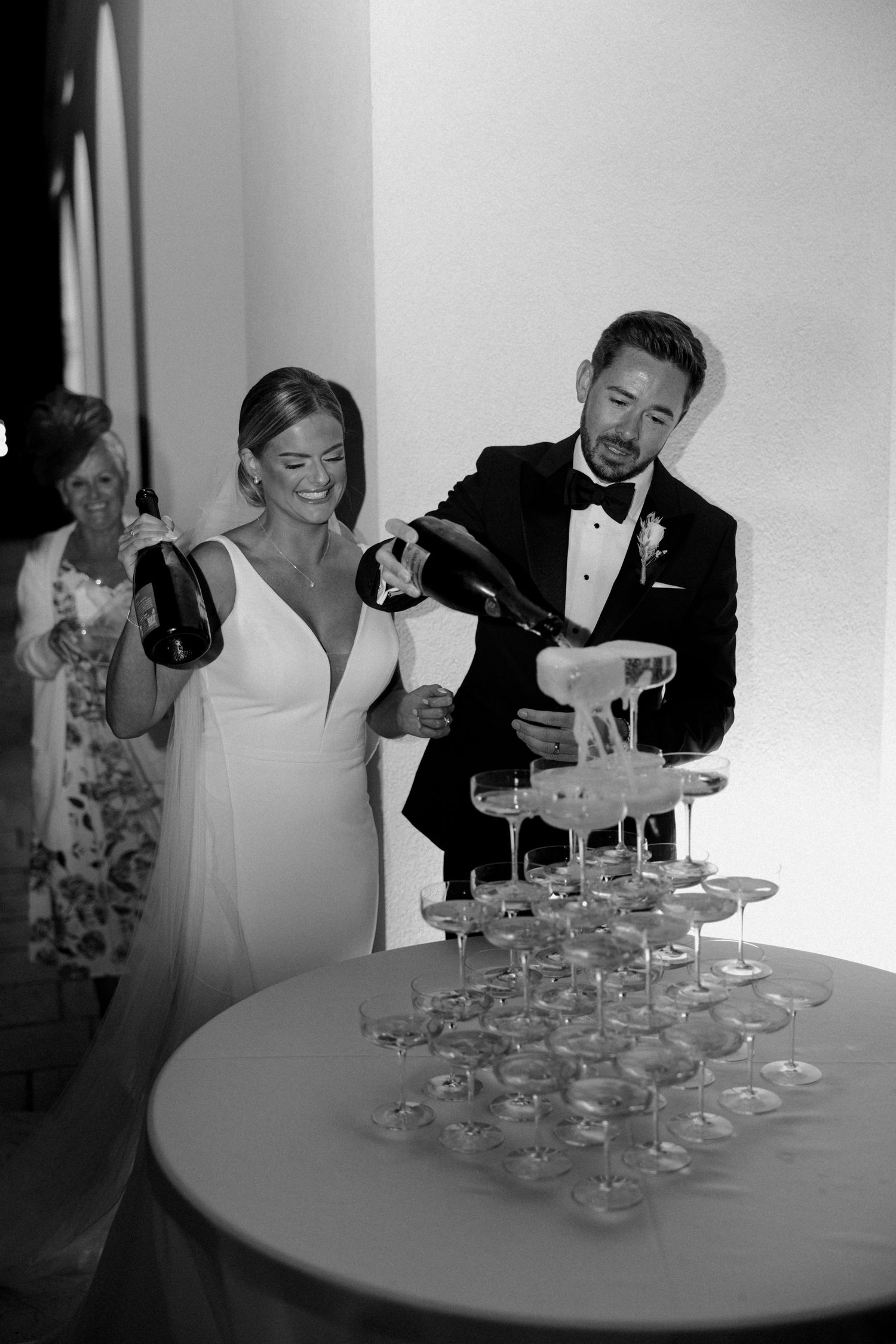 A couple pours champagne into a tower of elegant champagne glasses at their wedding celebration.