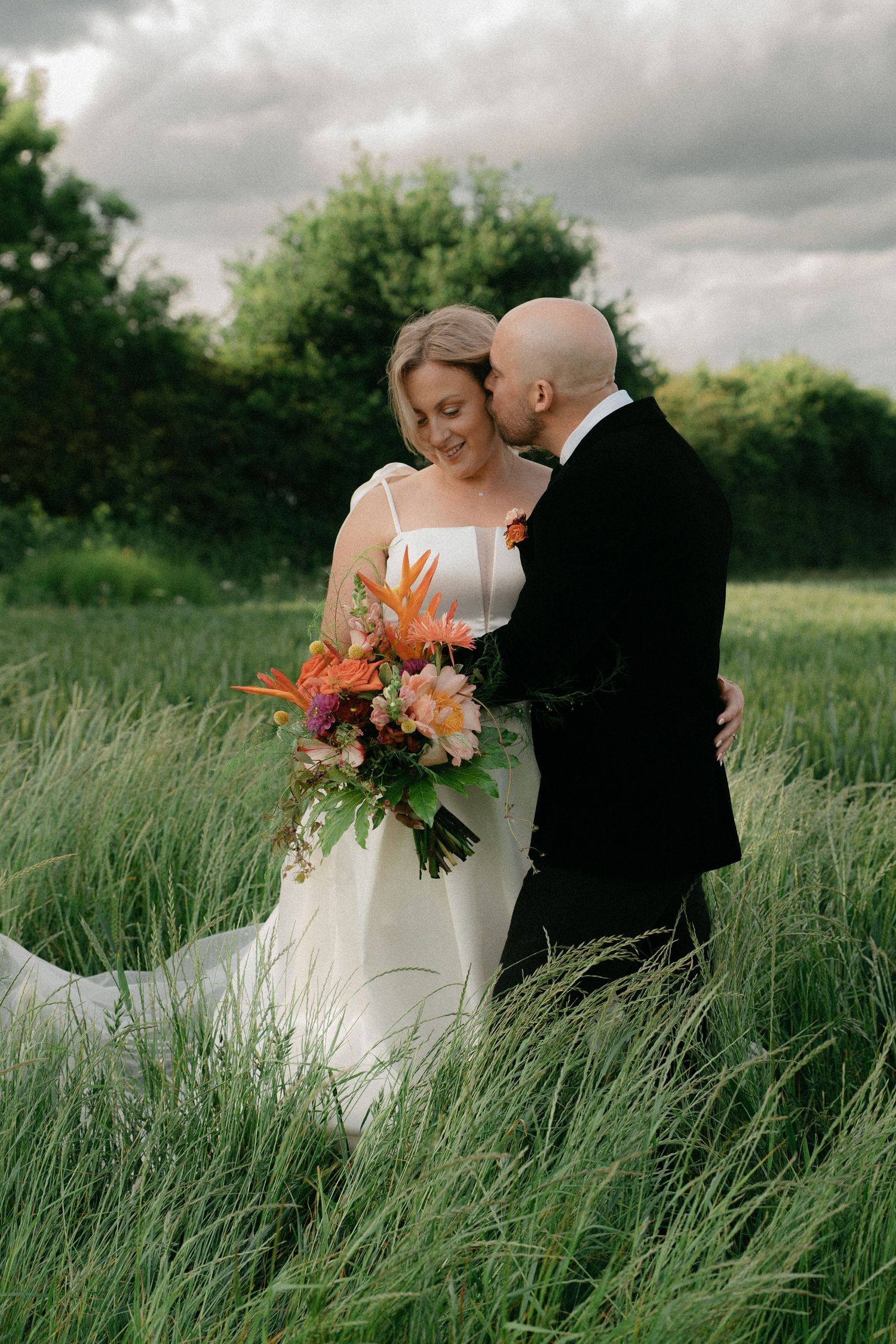 A romantic wedding photoshoot in a tall grass field with a beautiful orange and white bouquet.