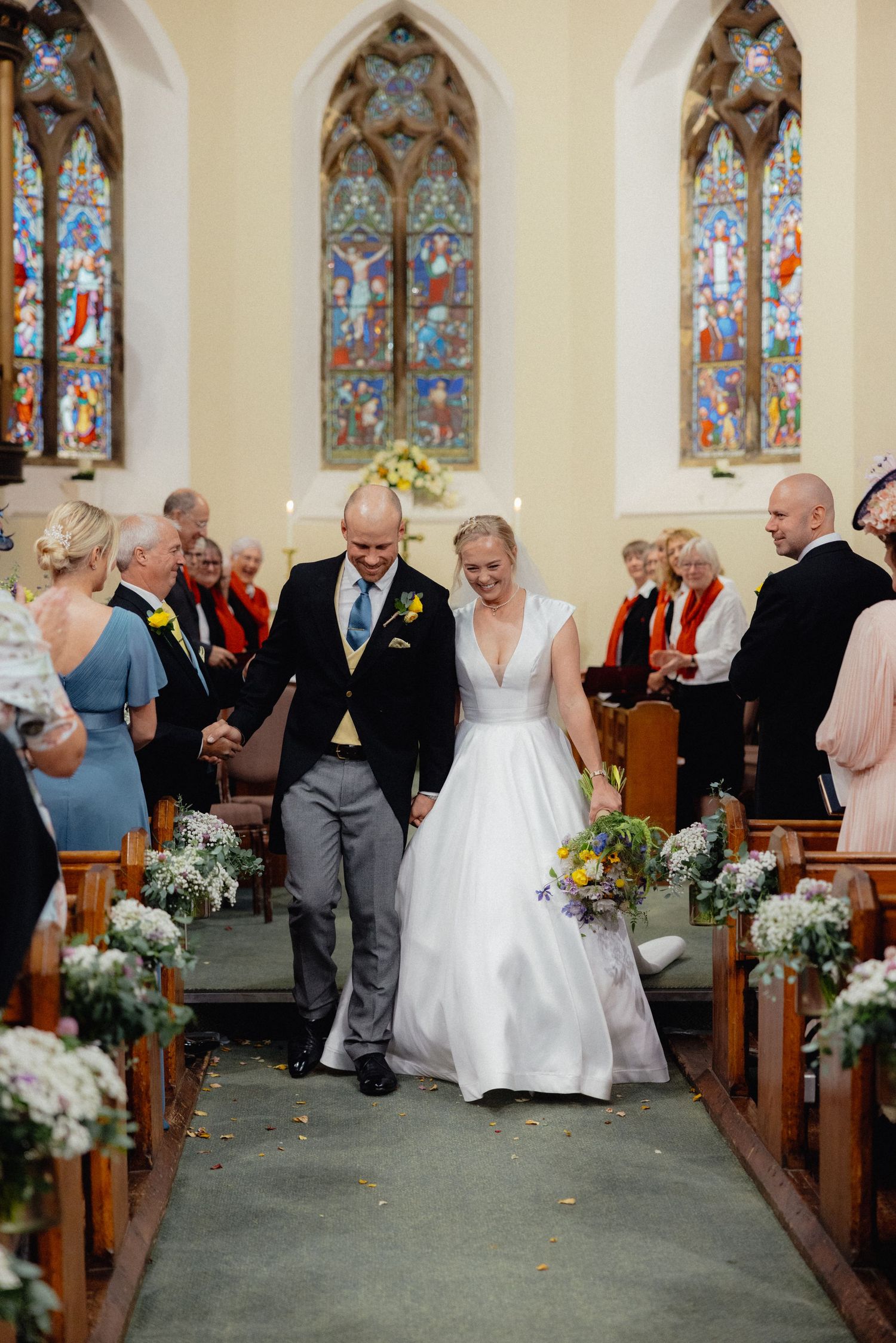 Wedding couple walking down church aisle decorated with white flowers and stained glass windows.
