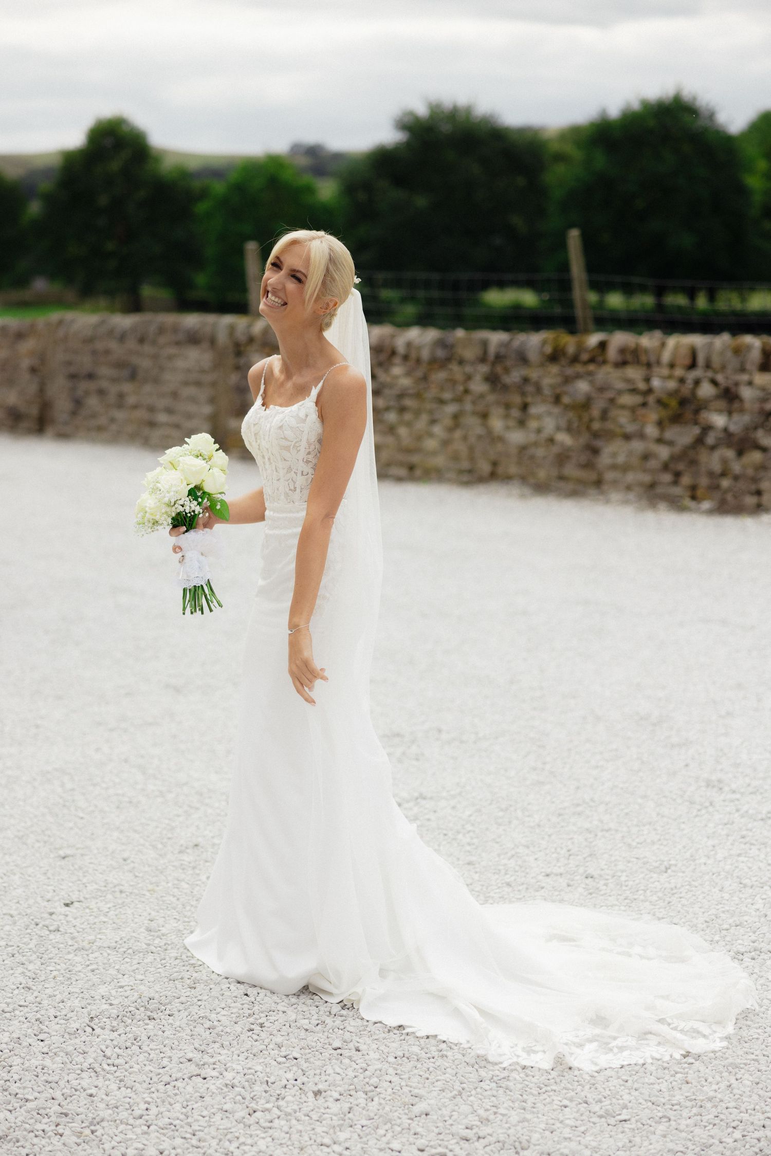 A bride in a white wedding gown with long train poses on a gravel path near stone walls.
