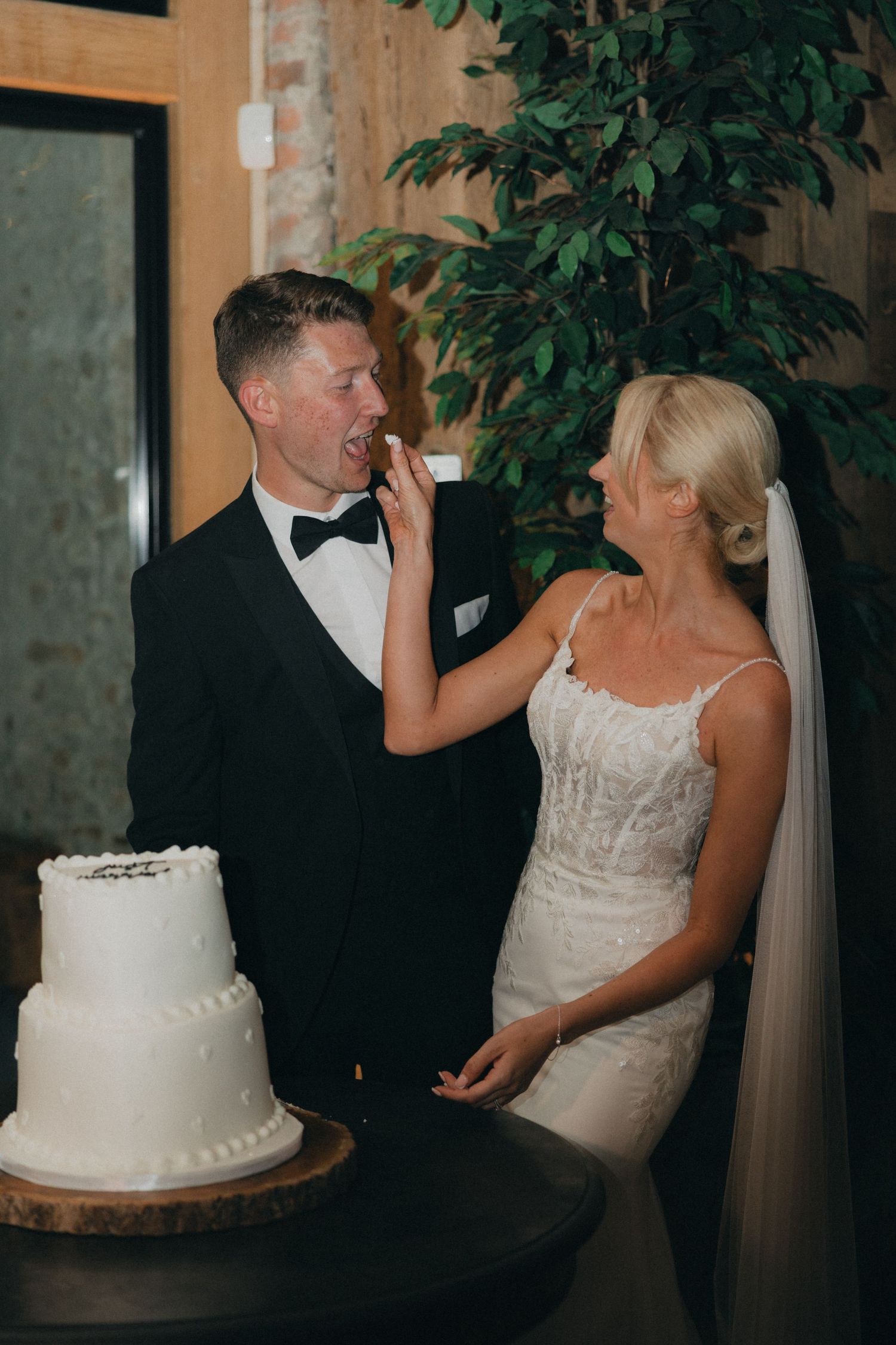 A playful moment as the bride feeds wedding cake to the groom beside their two-tier white cake.