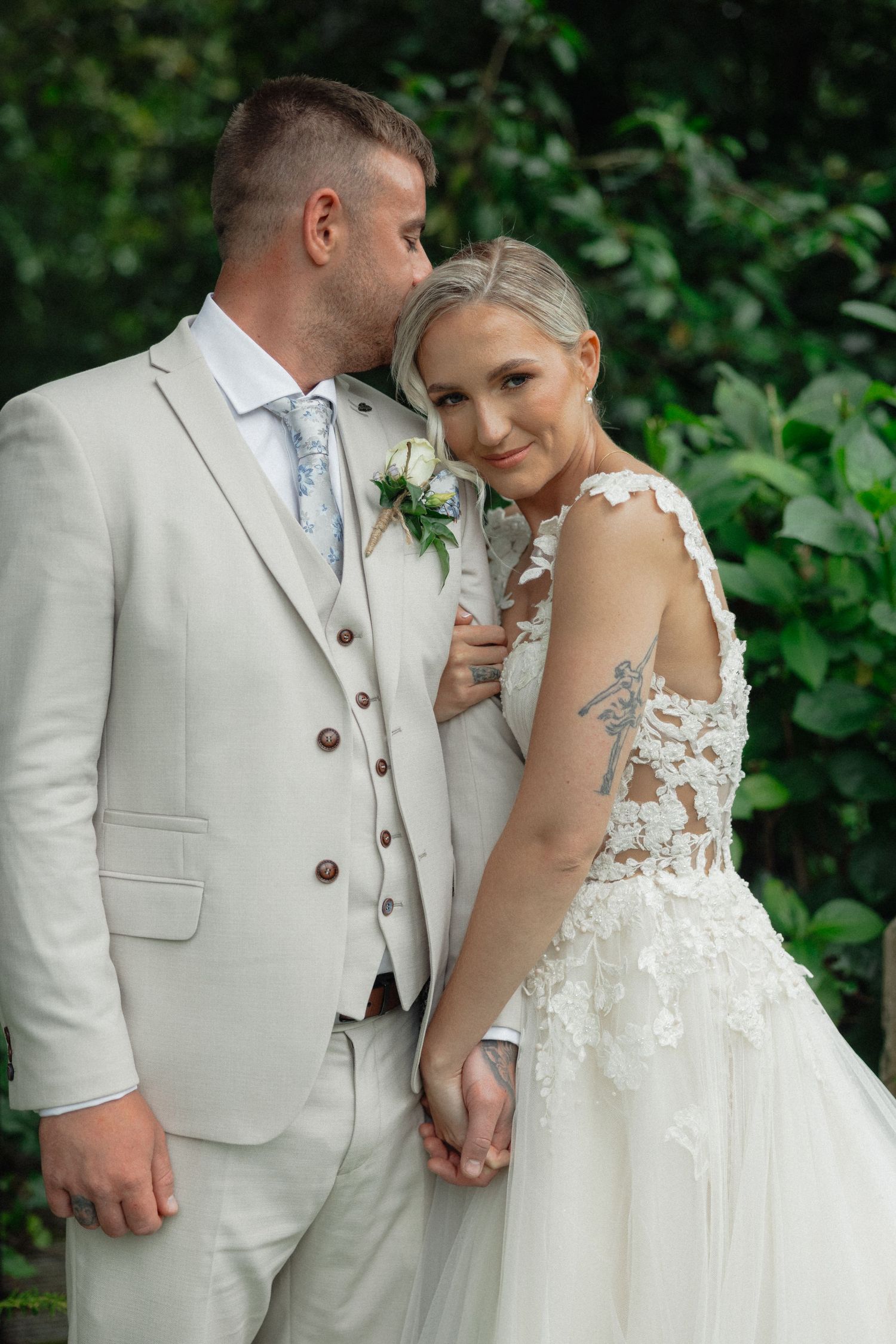 Romantic wedding couple in light-colored attire embracing in a garden setting.