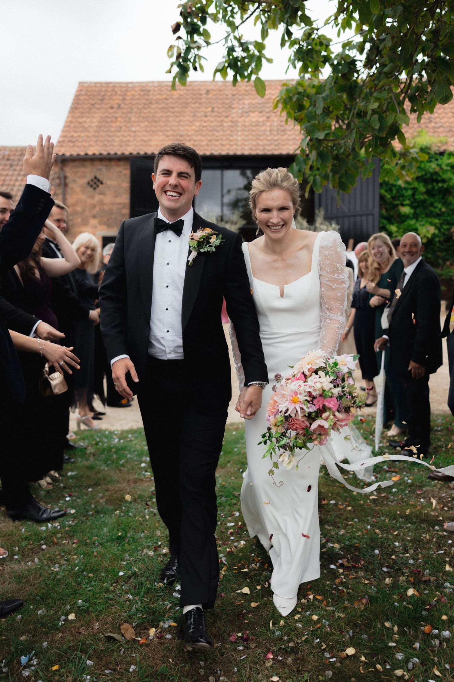 Newlyweds celebrating outdoors with guests on a lawn with scattered leaves.