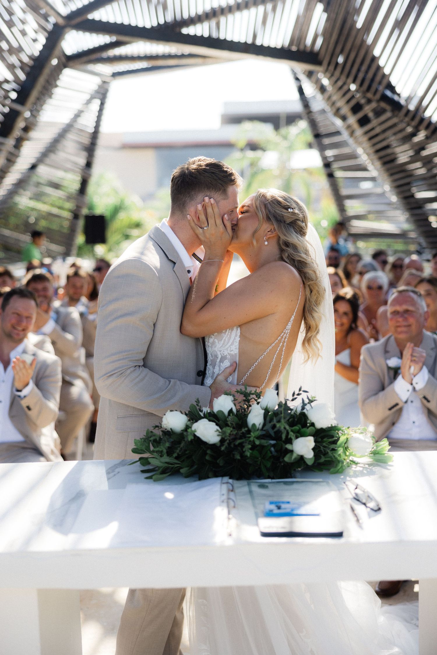 Newlyweds share first kiss at outdoor wedding ceremony while guests applaud.