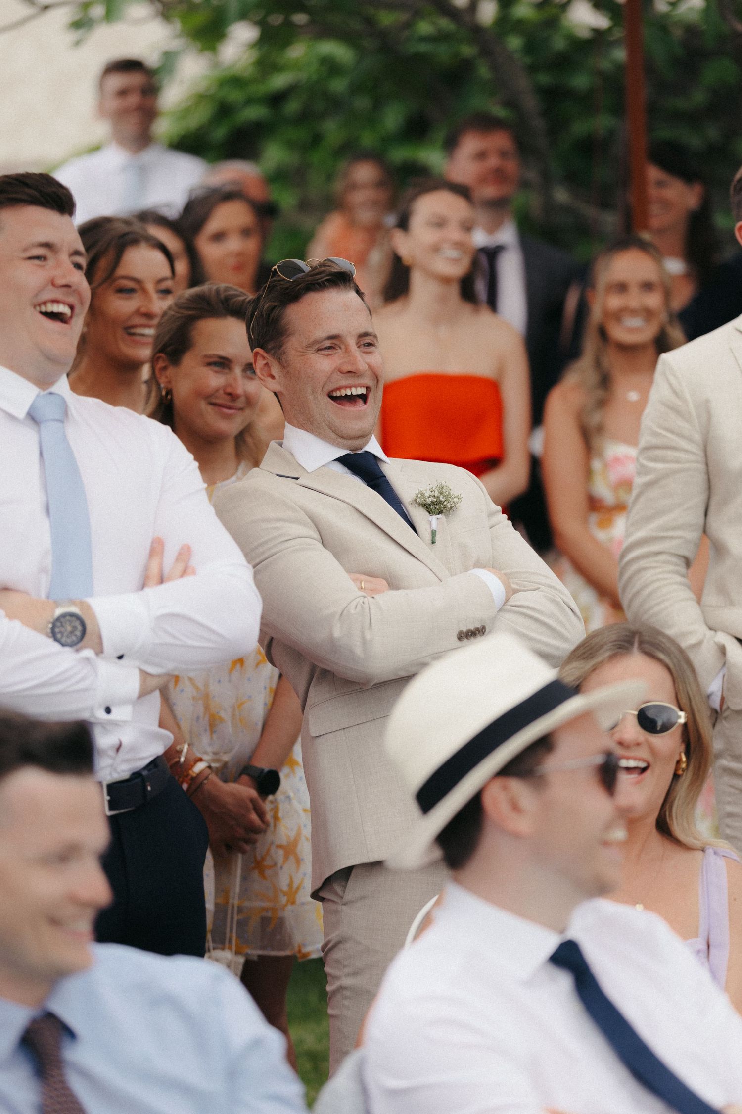 Wedding guests in light colored attire laugh joyfully during an outdoor celebration.