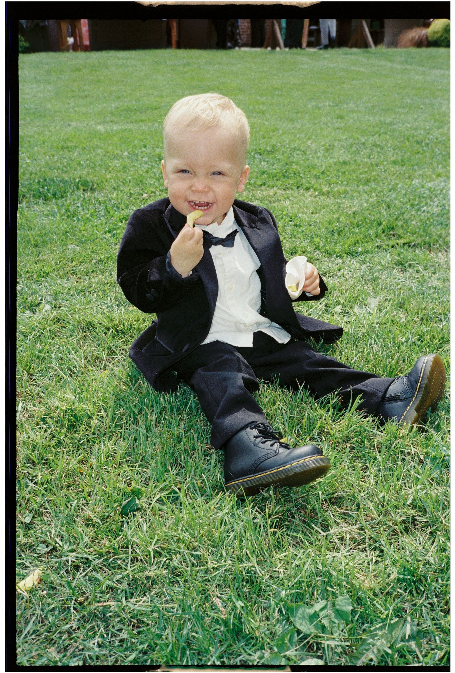 A young boy in formal attire sits playfully on grass during a special occasion.
