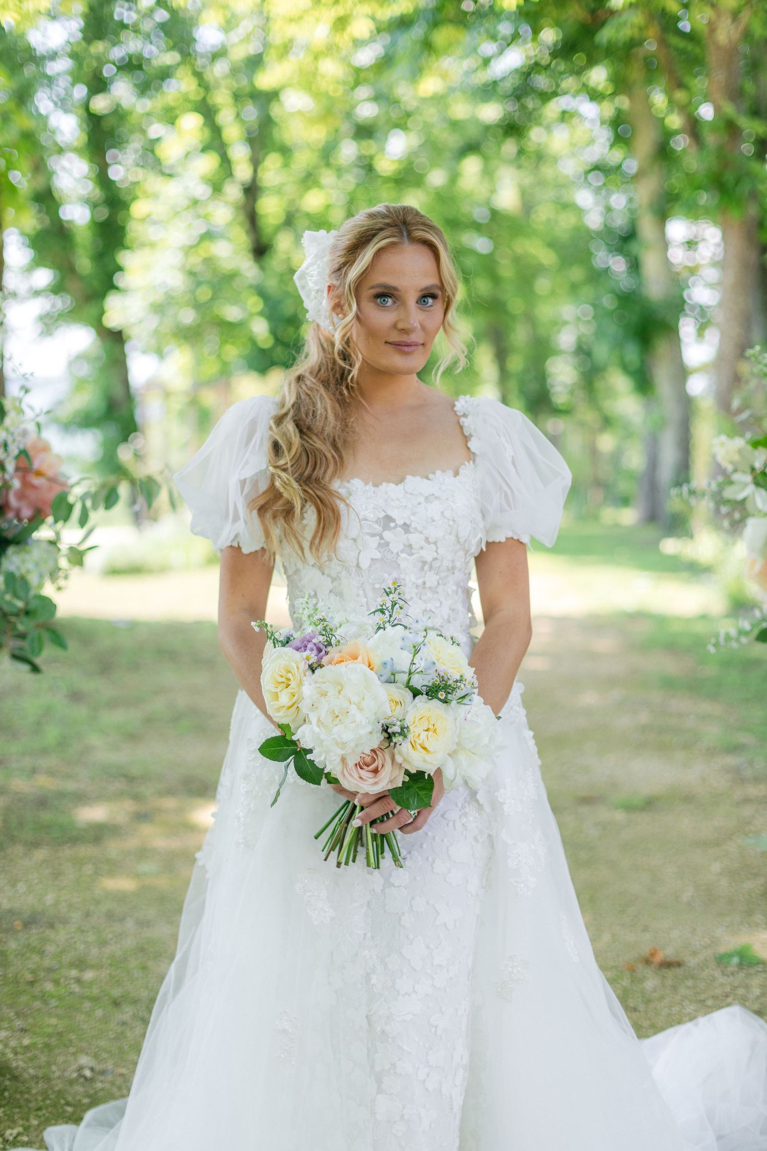 A bride in a white lace dress holds a bouquet of pastel flowers in a wooded setting.