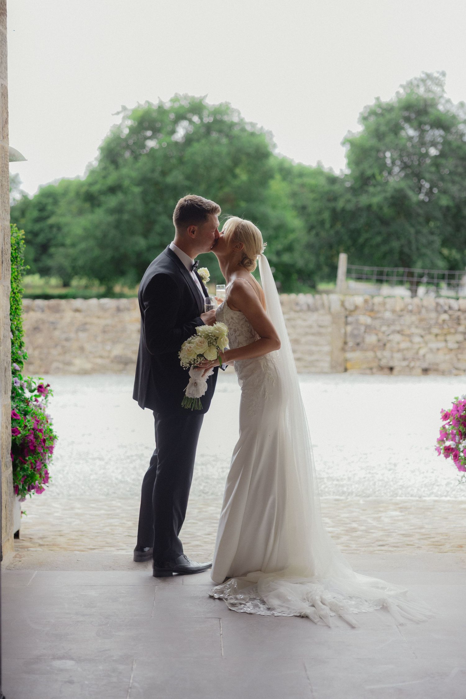 Wedding couple poses under covered porch with stone wall and lake backdrop.