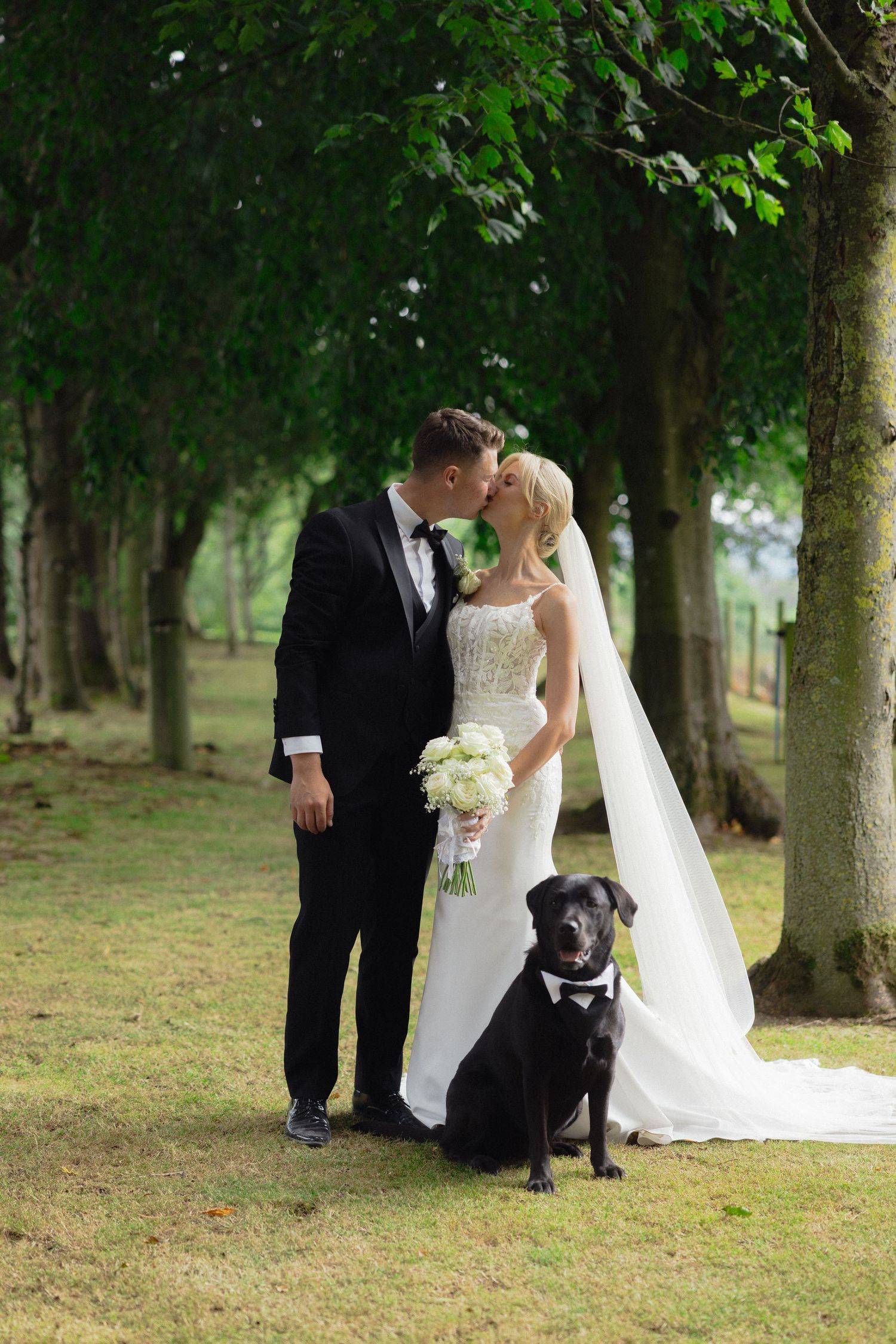 Newlyweds and their black dog share a moment in tree-lined garden setting.