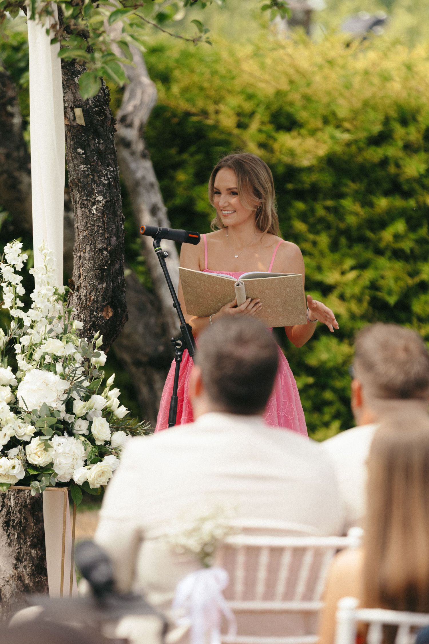 A speaker in a pink dress reads from a book during an outdoor wedding ceremony.