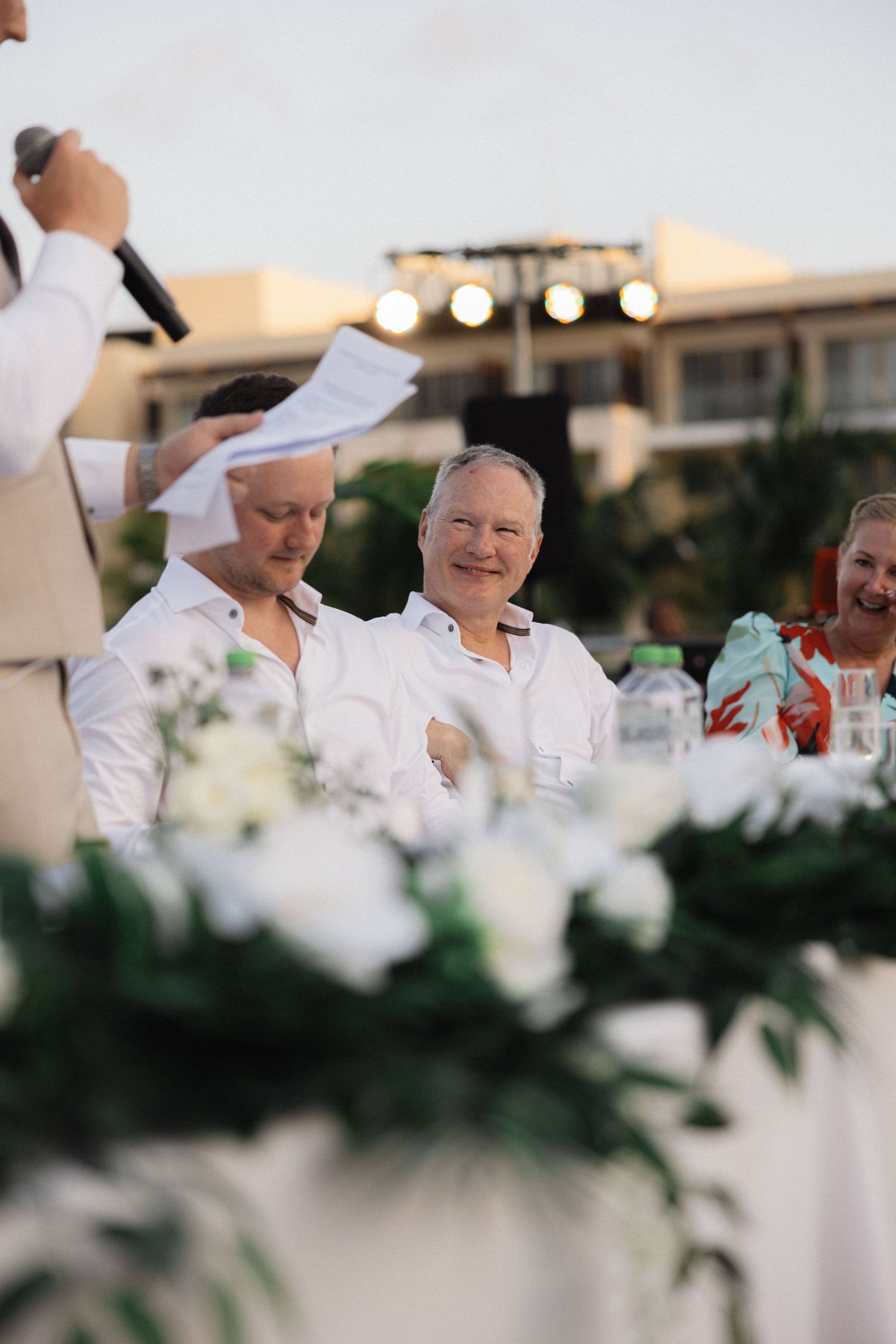 Wedding reception speech moment at an outdoor venue with white floral decorations.