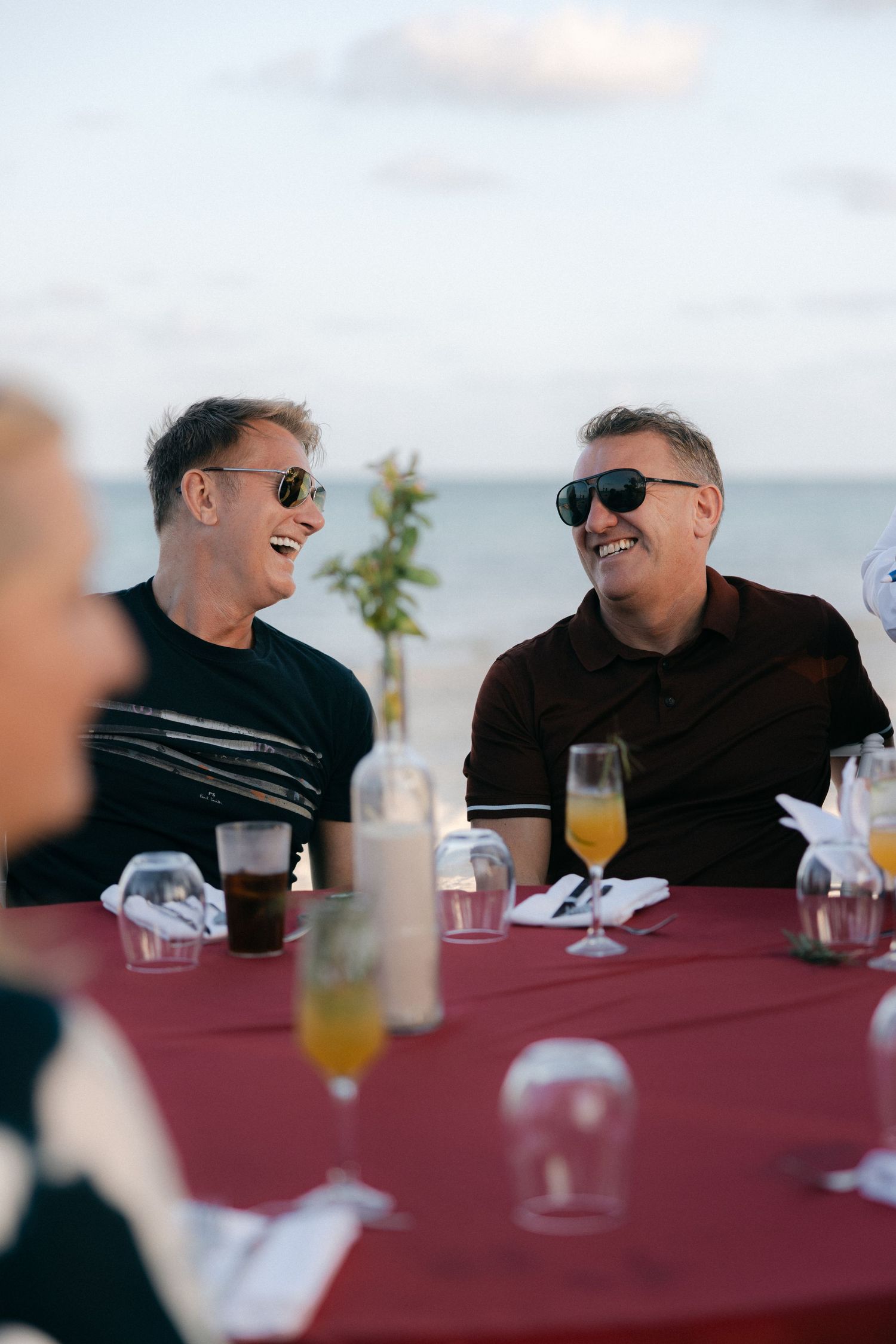 Two guests sharing laughs at outdoor dining table with drinks.