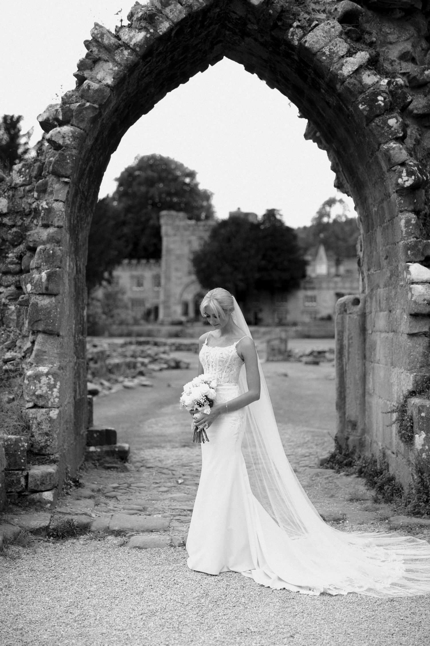 Black and white portrait of bride with flowing veil under stone archway.