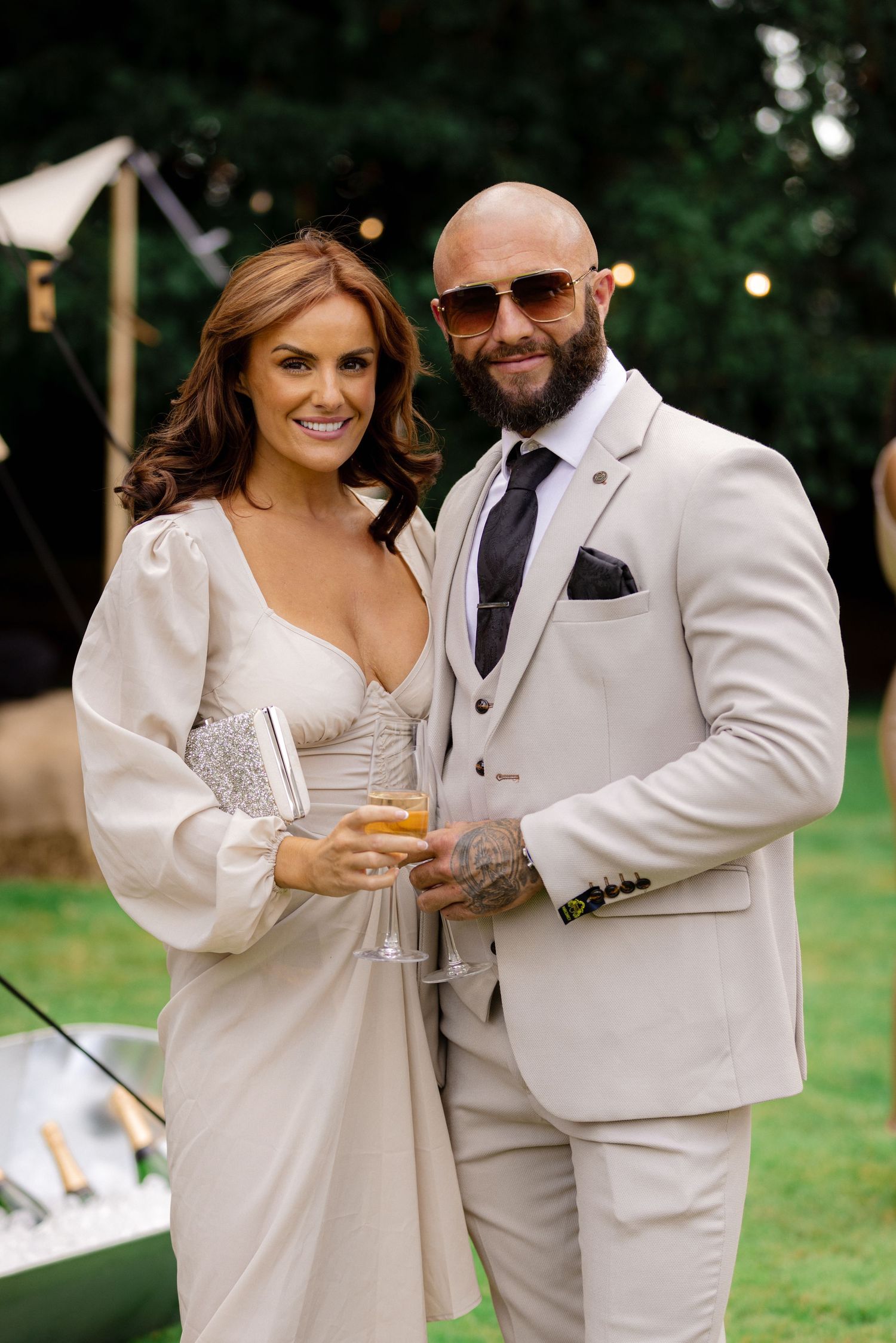 A stylish couple in coordinating cream-colored wedding attire toasting with champagne glasses outdoors.