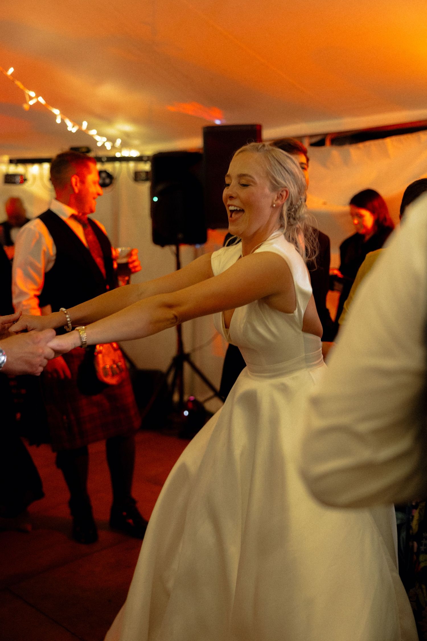 Bride in white dress dancing joyfully at wedding reception with orange ambient lighting.