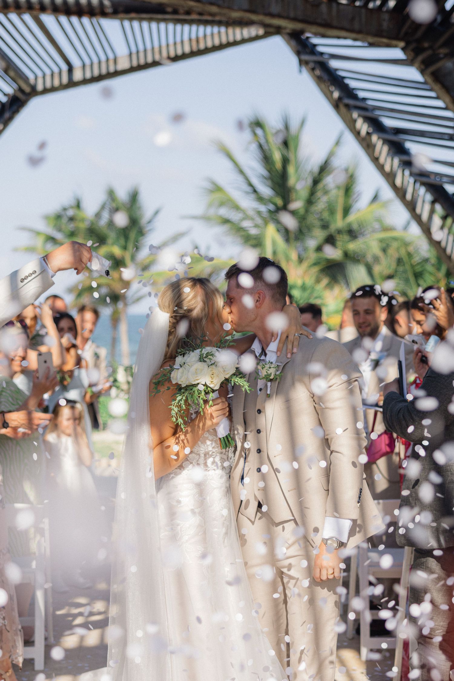 Wedding party celebrates with confetti under a covered pavilion surrounded by tropical palm trees.