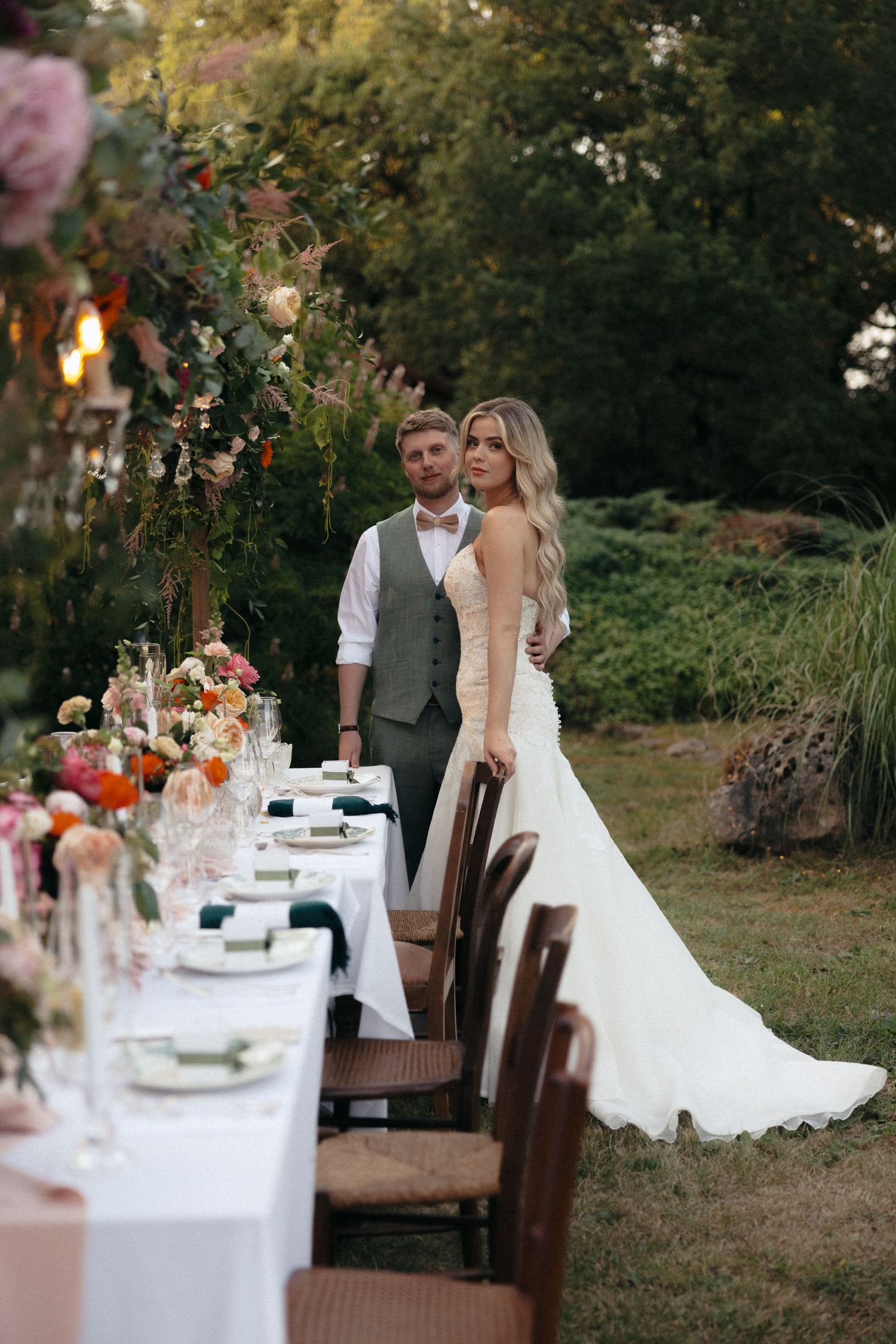 A couple poses by an elegant outdoor wedding table setting decorated with flowers at sunset.
