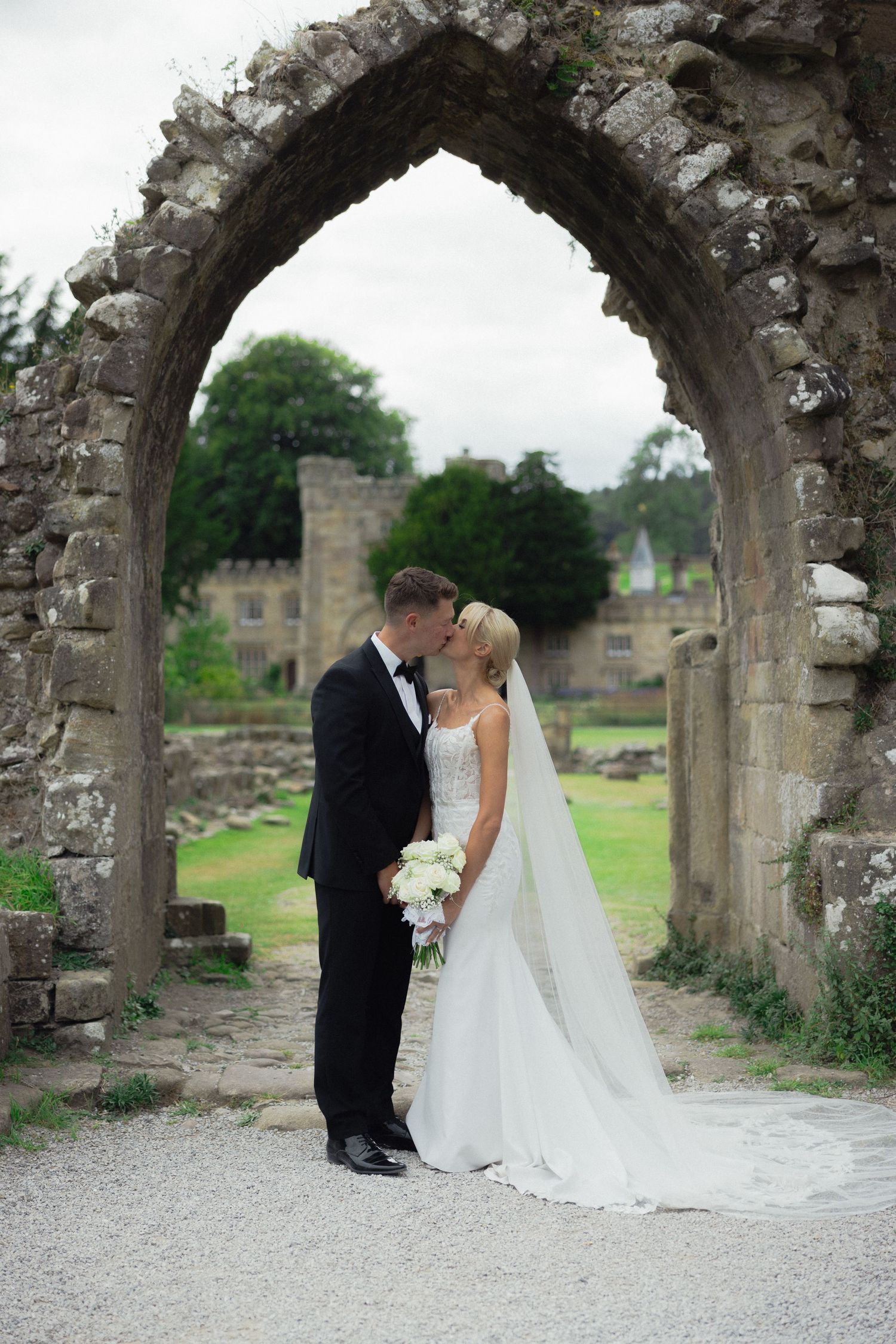 A romantic wedding photo under a stone archway at a historic ruins site.