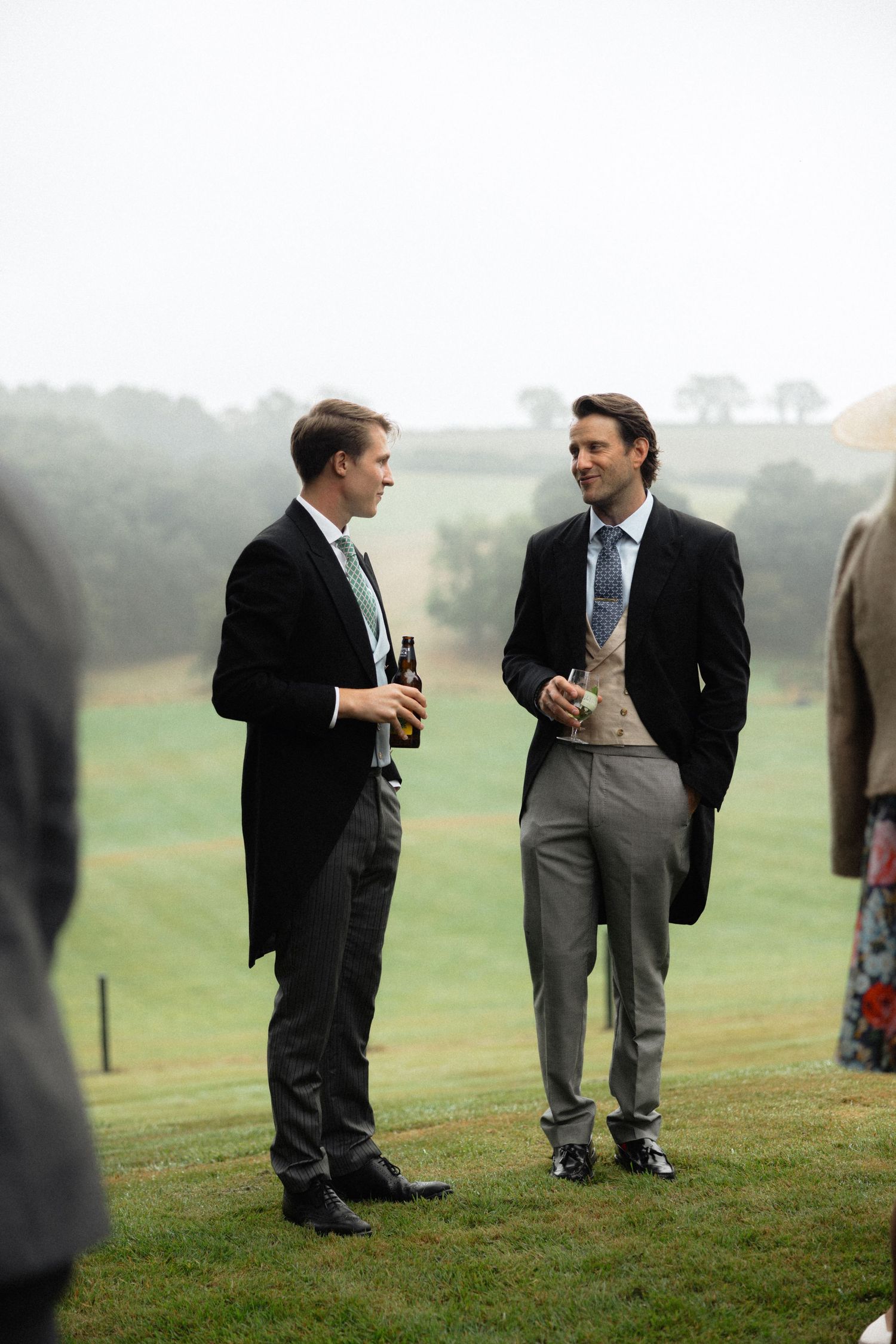 Wedding guests in formal attire having conversation on grassy hillside.