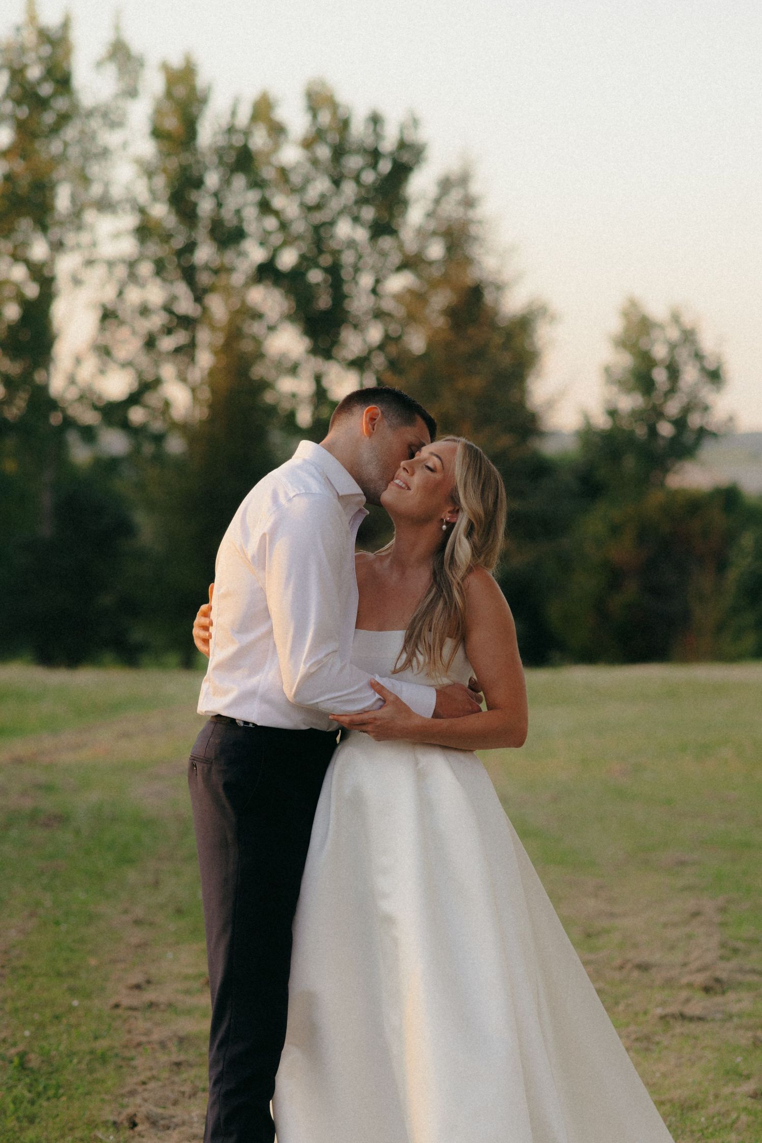 A romantic couple embraces in an outdoor field during golden hour wearing wedding attire.