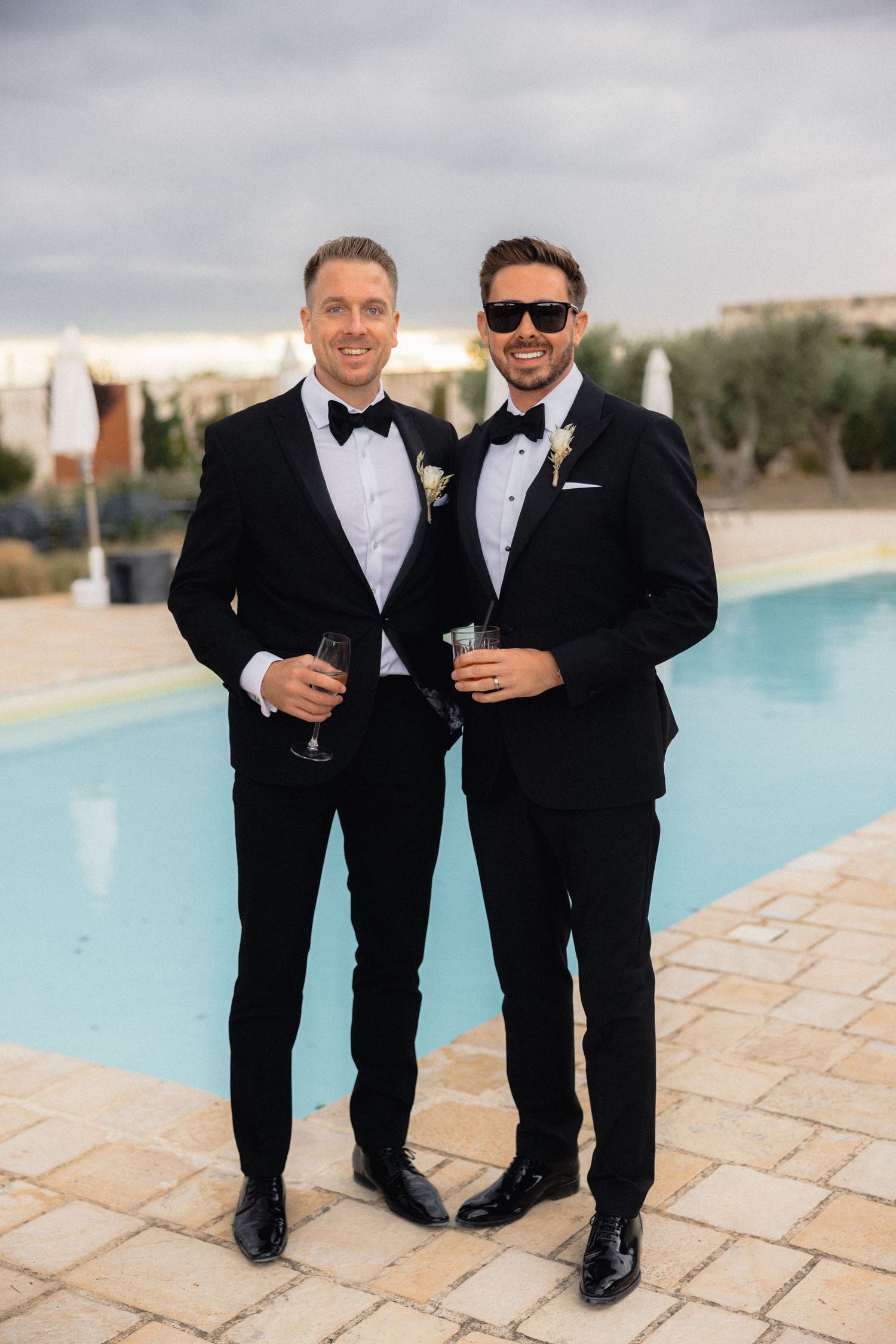 Groomsmen in black tuxedos sharing drinks and laughs by a swimming pool.