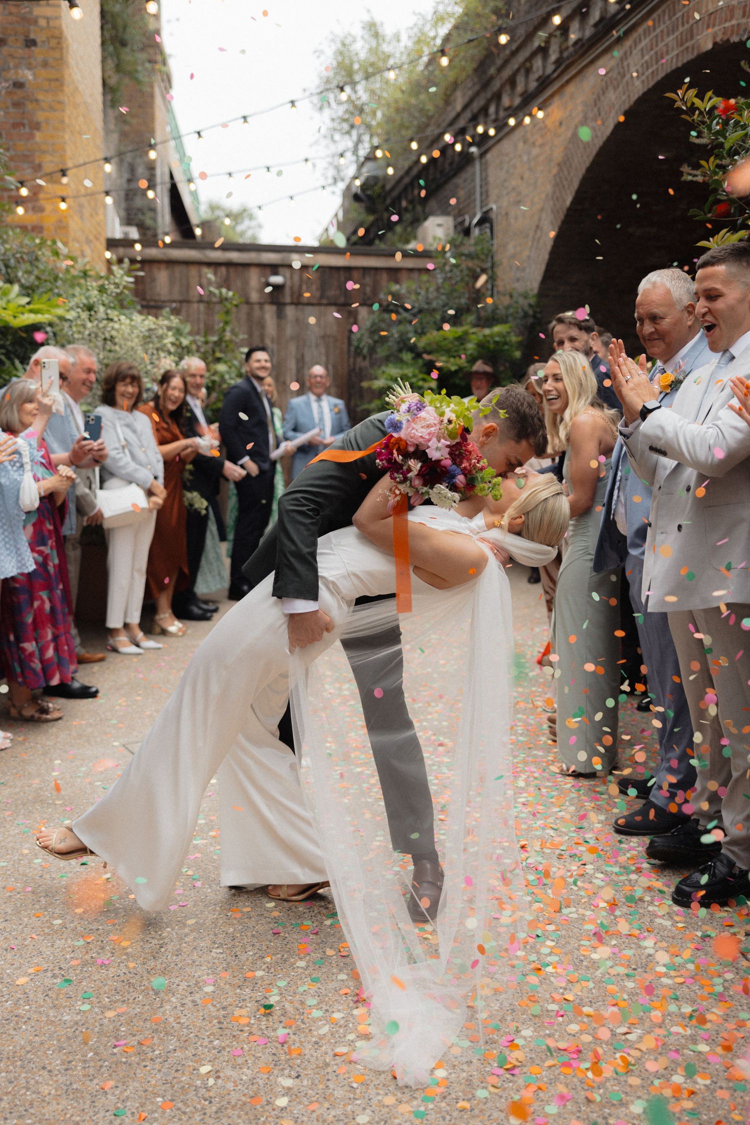Wedding couple celebrates under shower of colorful confetti with guests in brick archway venue.