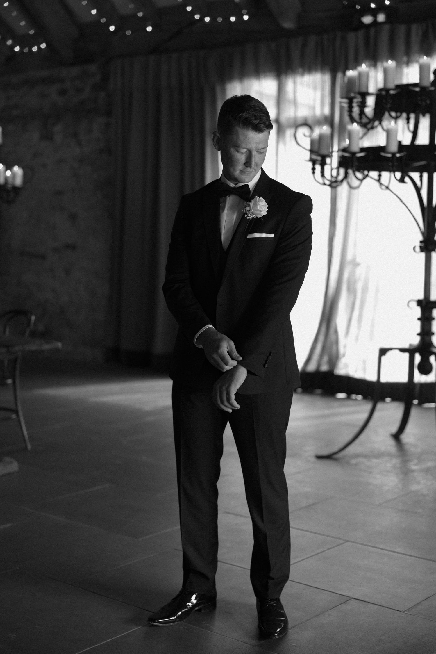 Formal portrait of groom in black tuxedo standing in elegant venue with string lights.