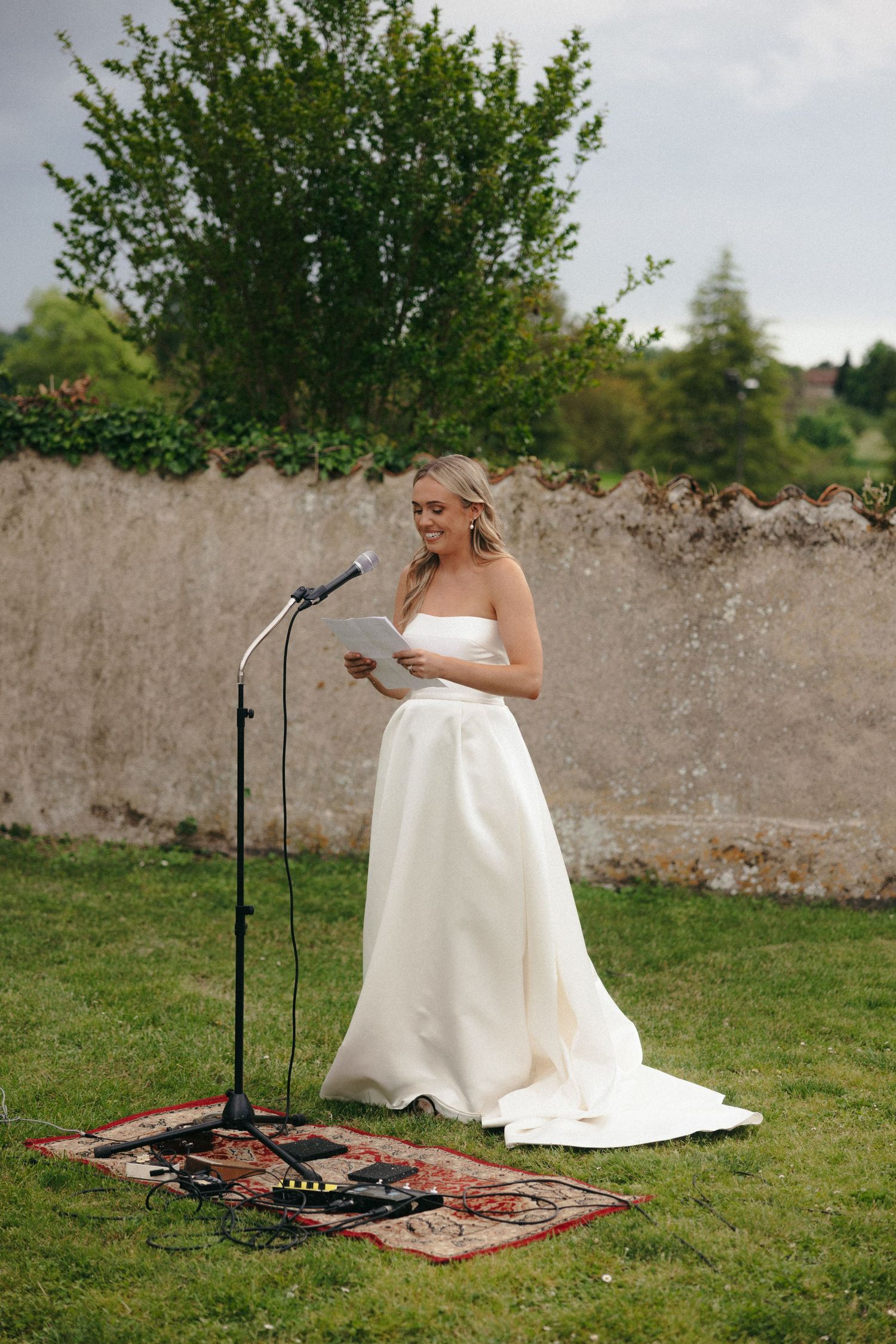 Bride in white dress standing near microphone equipment on grass with stone wall backdrop.