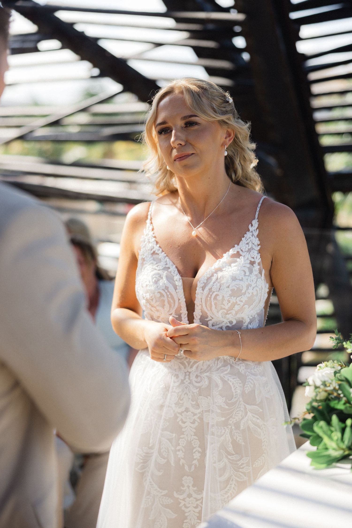 Bride in white lace dress with delicate straps listens intently during outdoor ceremony.