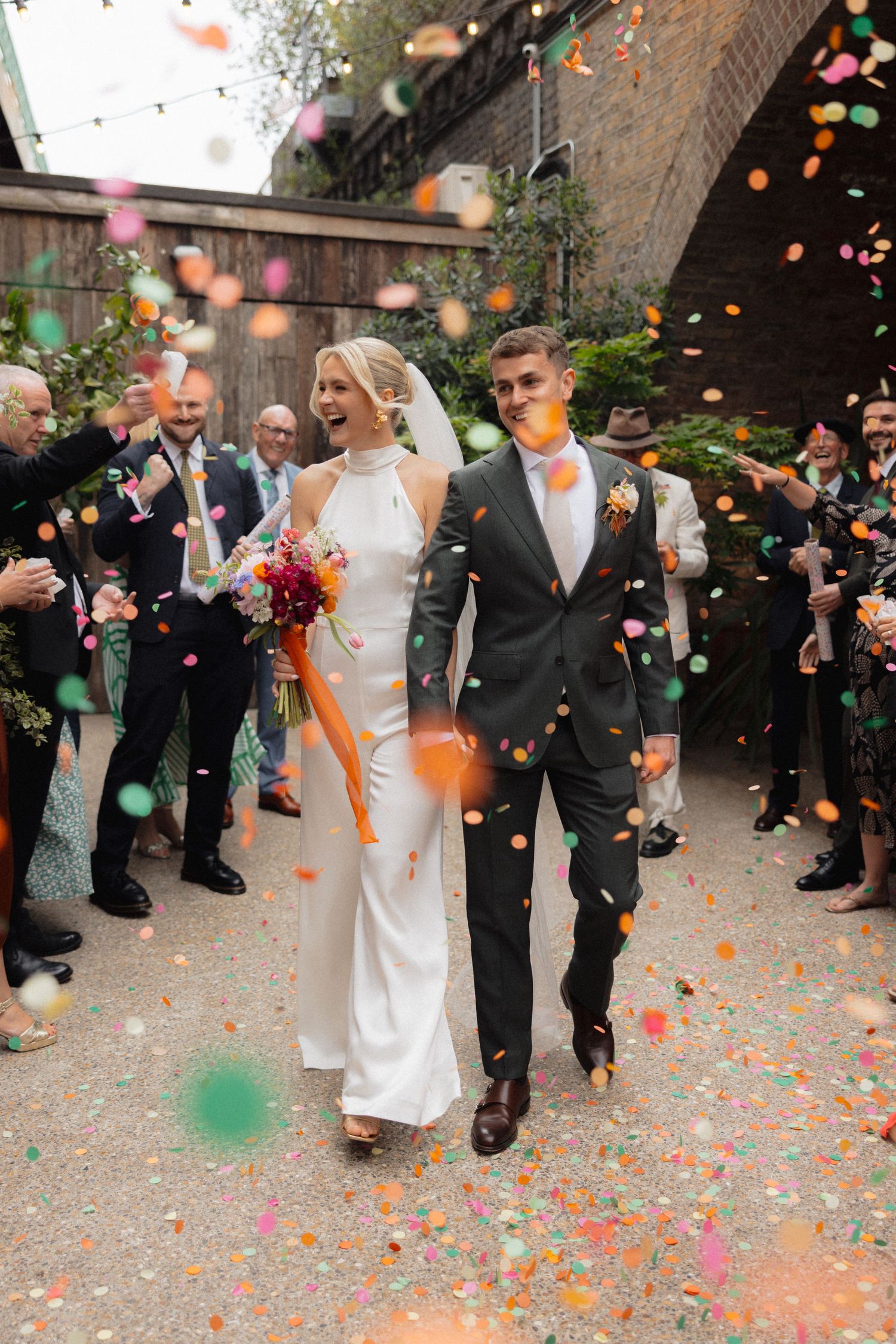 A wedding couple walks through colorful confetti shower with guests celebrating in the background.