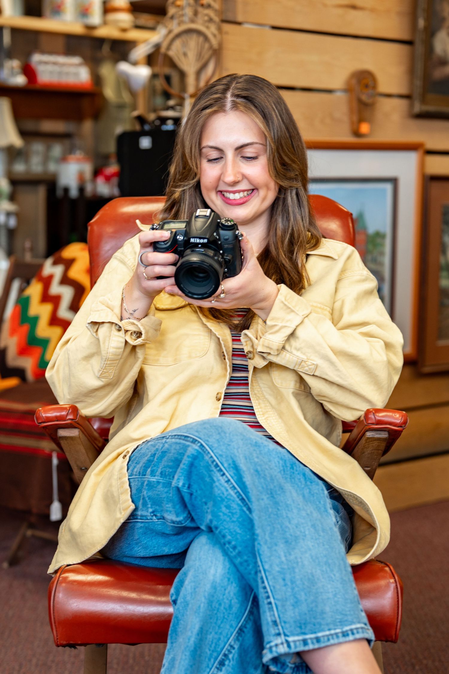 Smiling woman in yellow jacket and jeans sitting in red leather chair, reviewing photos on Nikon camera in vintage shop.