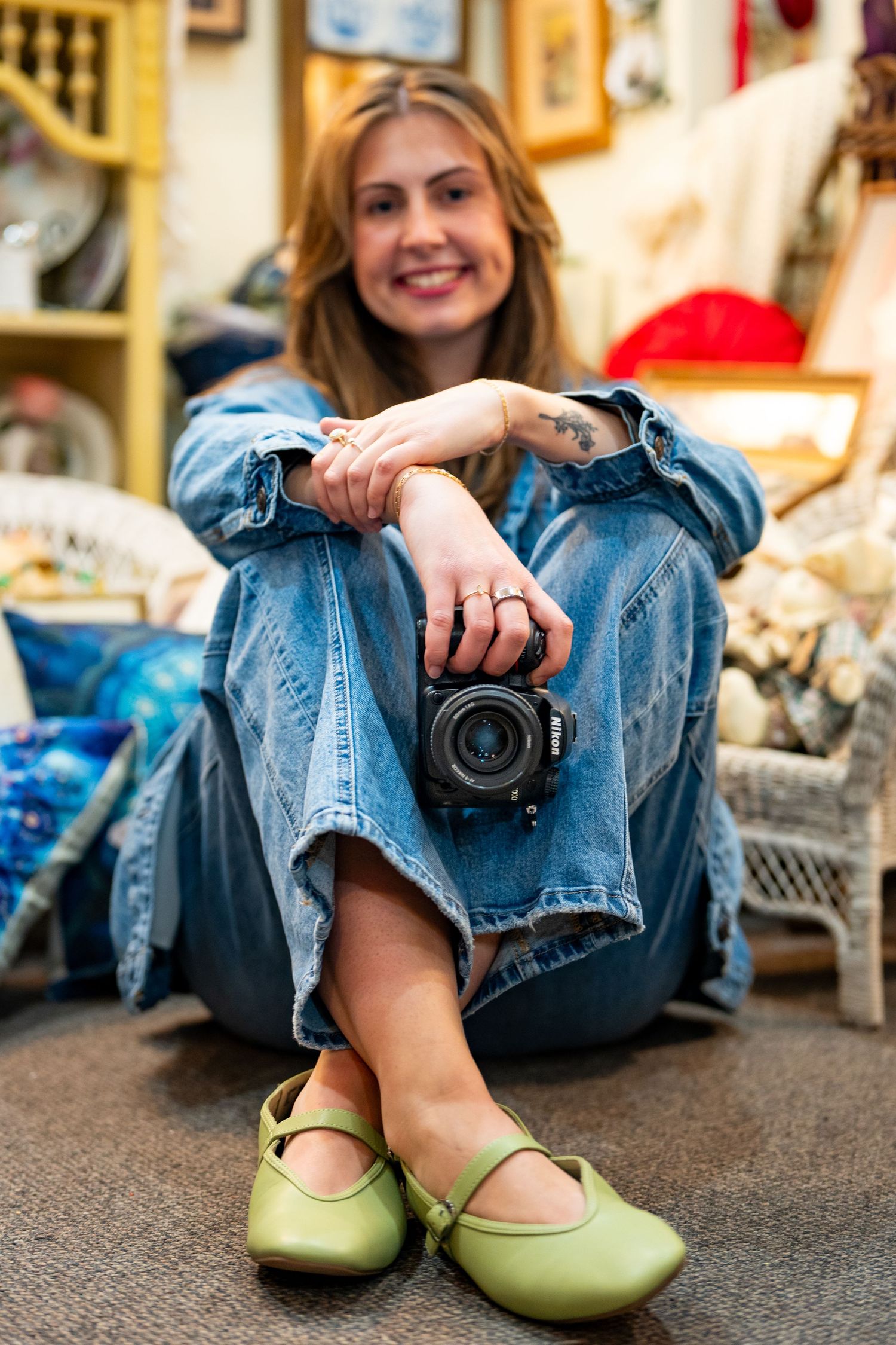 Smiling woman in denim outfit sitting on floor holding a camera, wearing green Mary Jane flats in a cozy vintage shop.