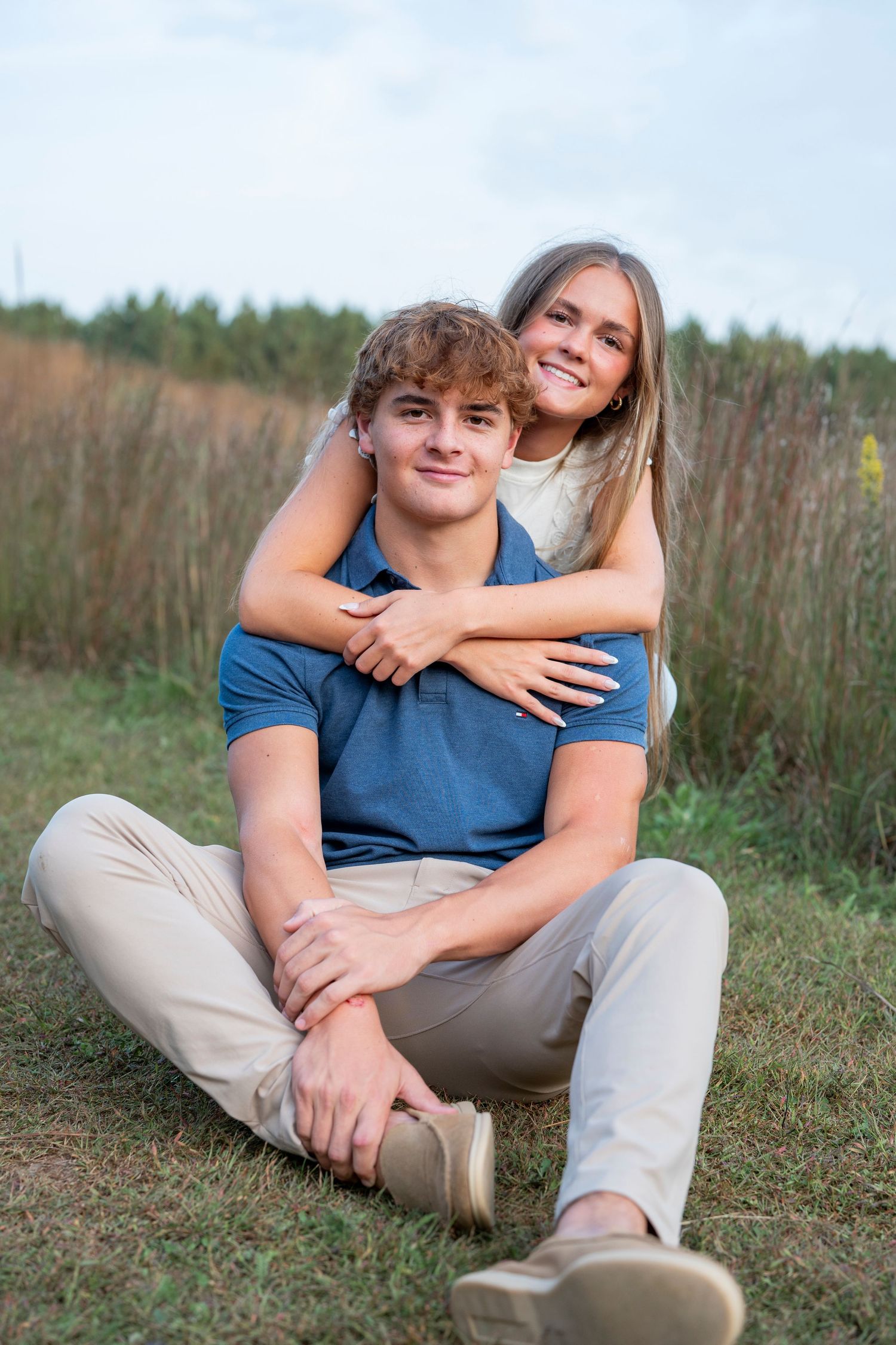 A couple sitting together in a field with tall grass and trees in the background.