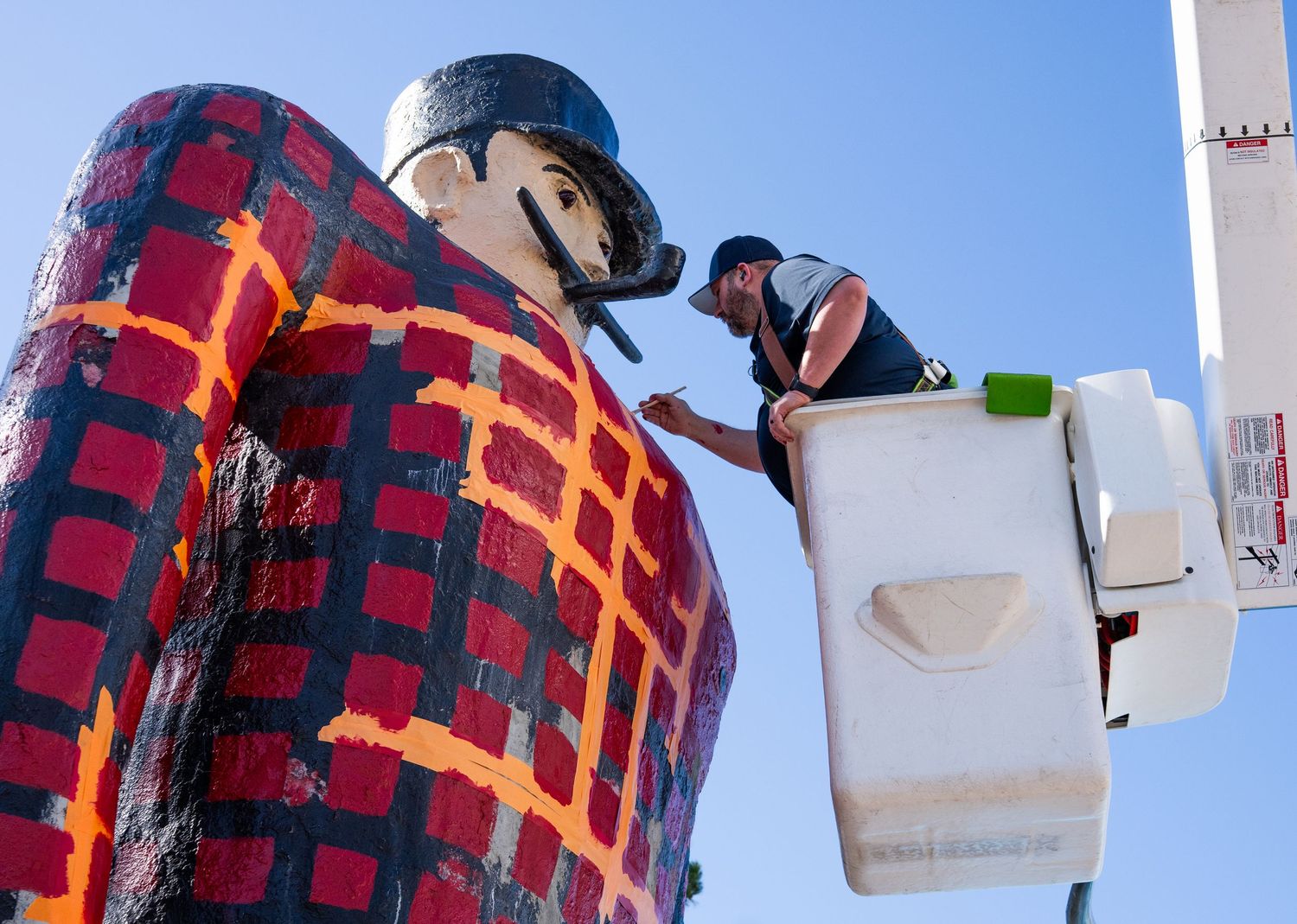 Artist painting details on a large Paul Bunyan statue wearing a red plaid shirt against a bright blue sky.