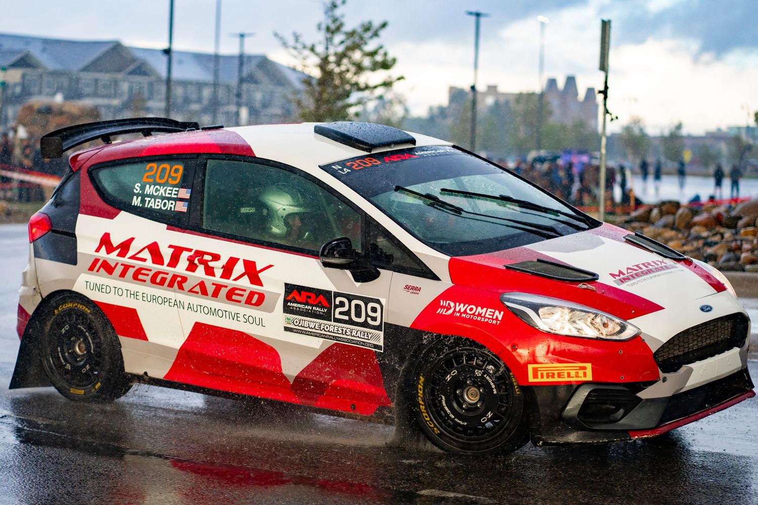 Red and white rally cars racing on wet asphalt track.
