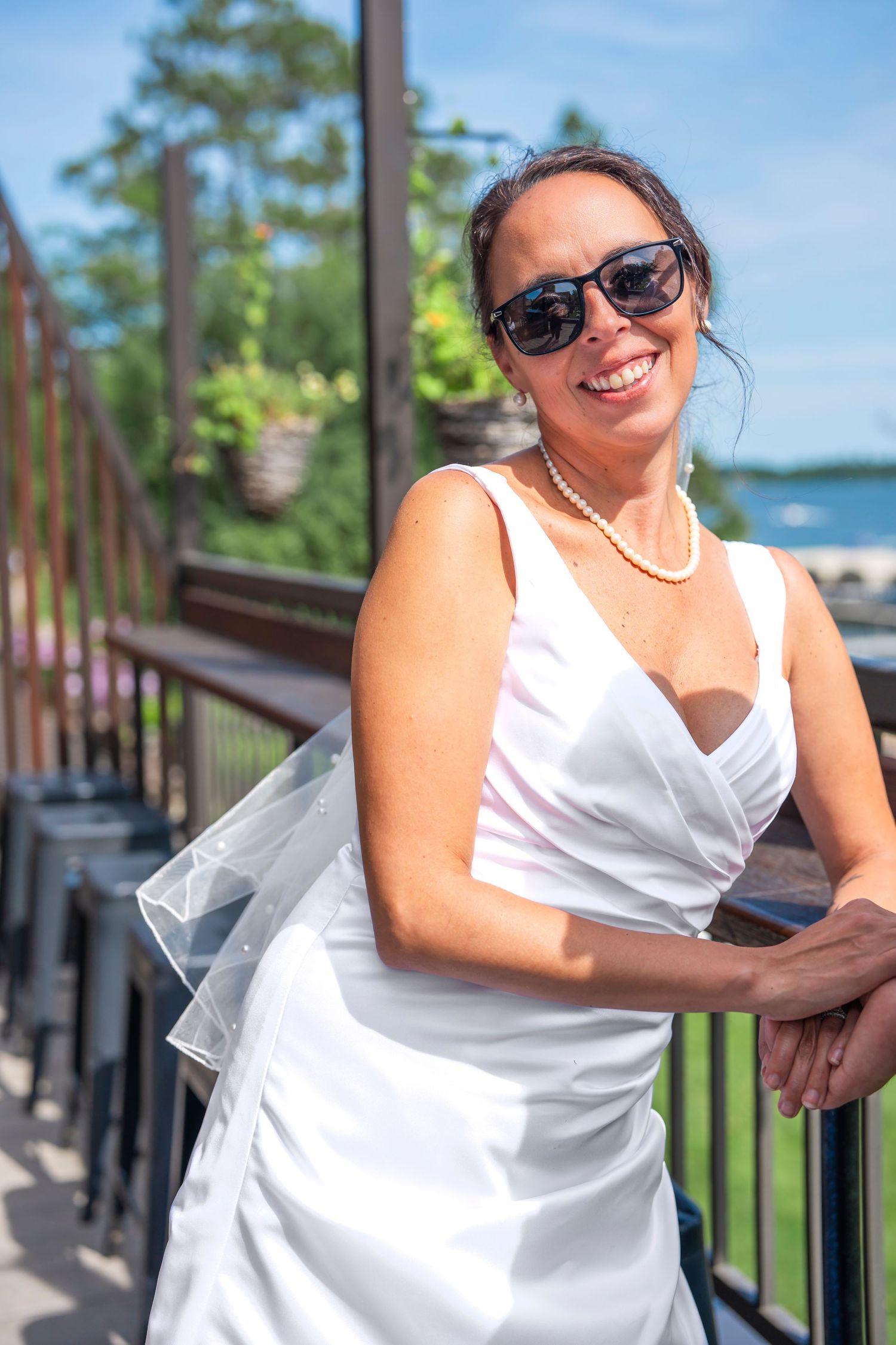 A bride in a white dress poses playfully on a wooden deck with water and trees visible behind.