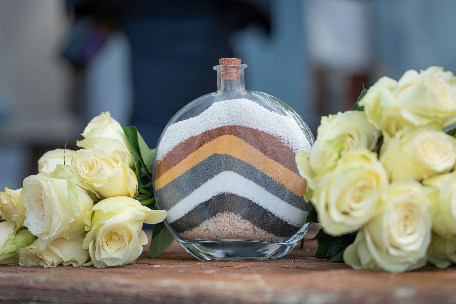 Decorative glass bottle with layered sand between white roses on wooden surface.