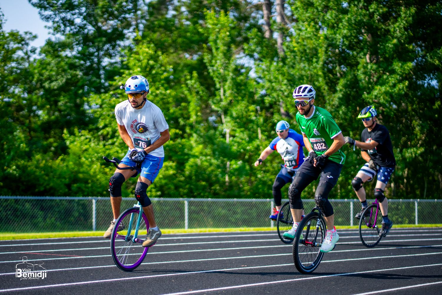 Athletes riding unicycles compete on blue running track during summer event.